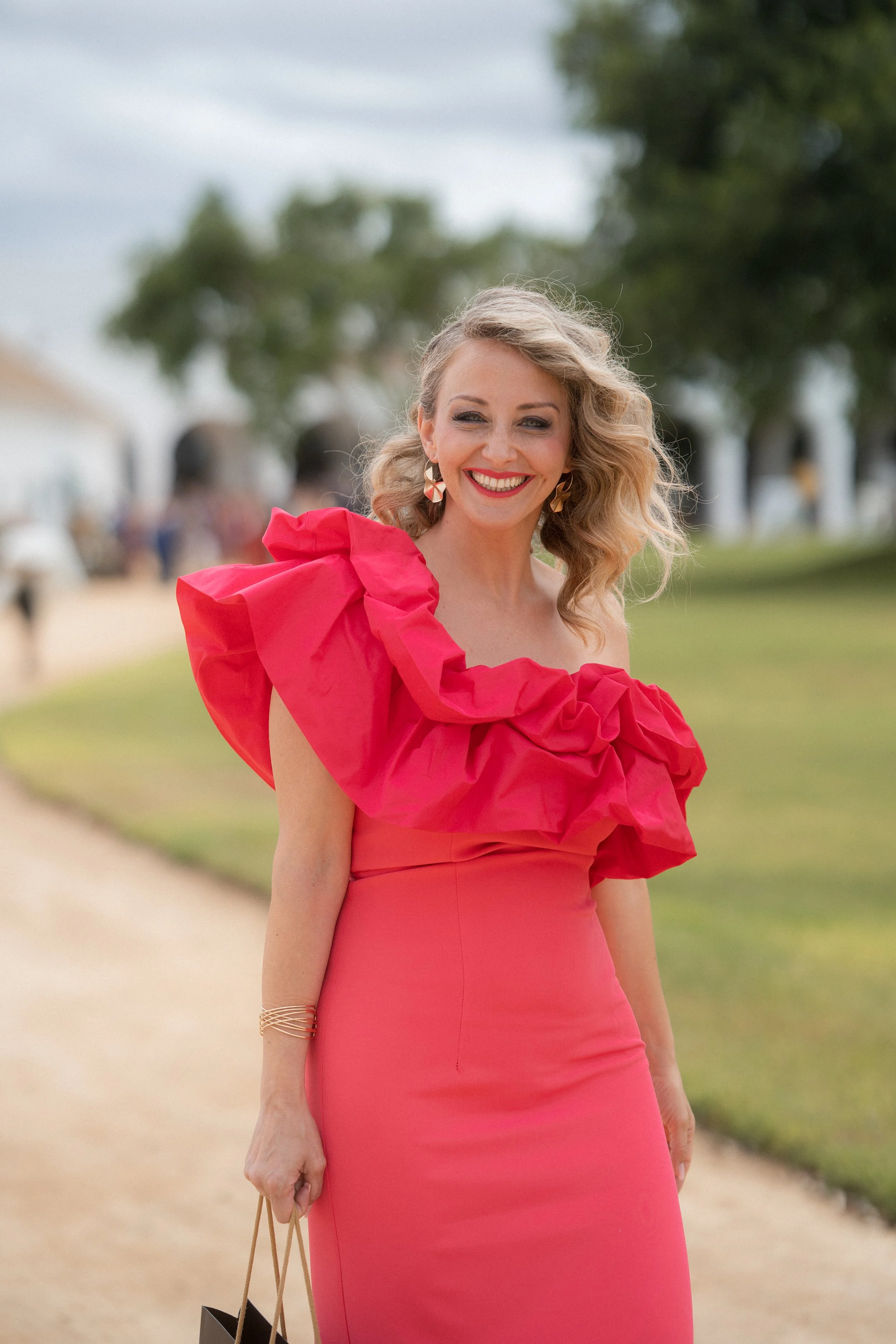 Mujer sonriendo con vestido largo y rosa, con un gran lazo en el hombro, en un entorno al aire libre con árboles y caminos.