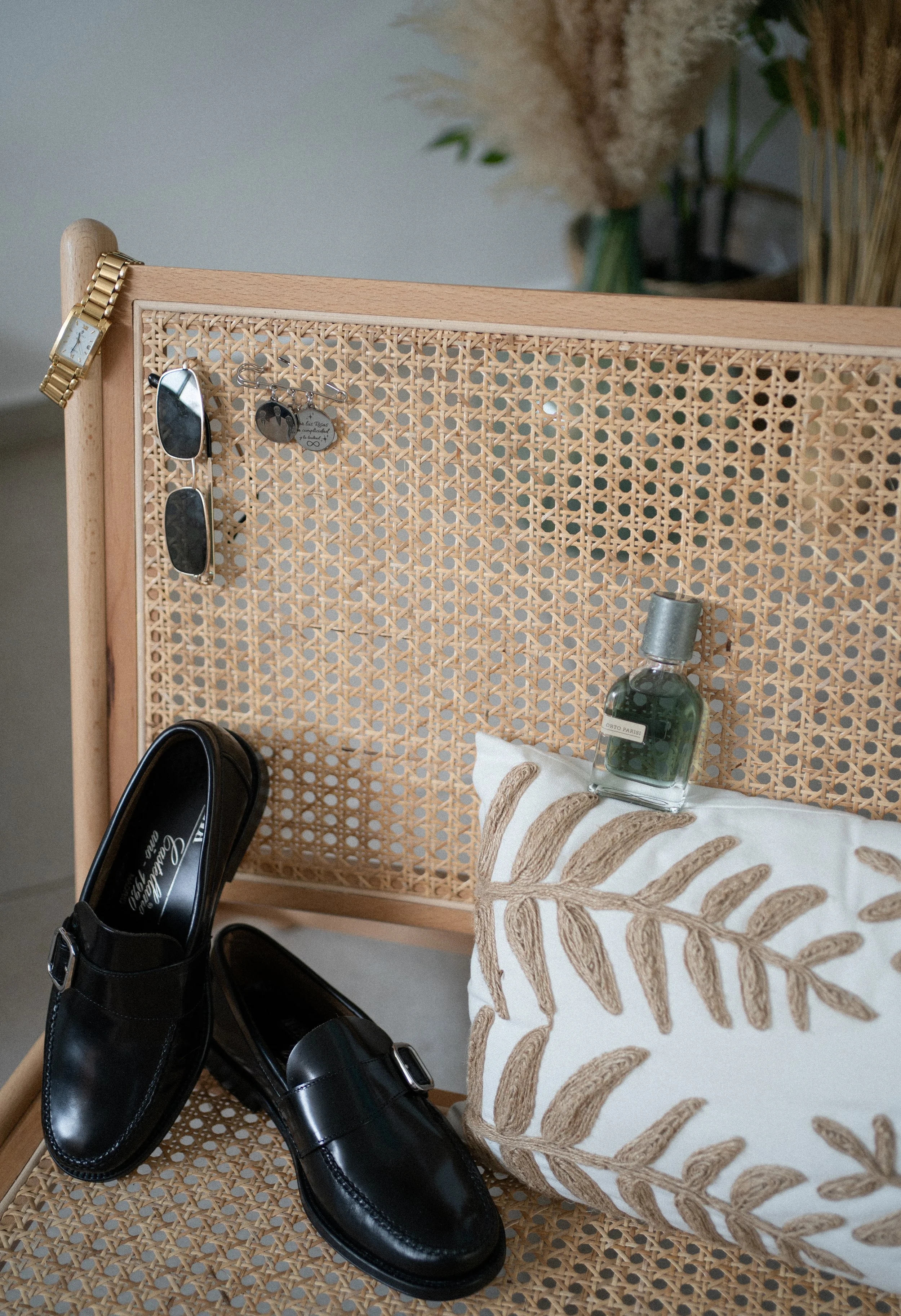 Zapatos negros de charol, lentes de sol, pendientes, botella de perfume, almohada decorativa con diseño de hojas y fondo de mimbre con piso de cerámica.