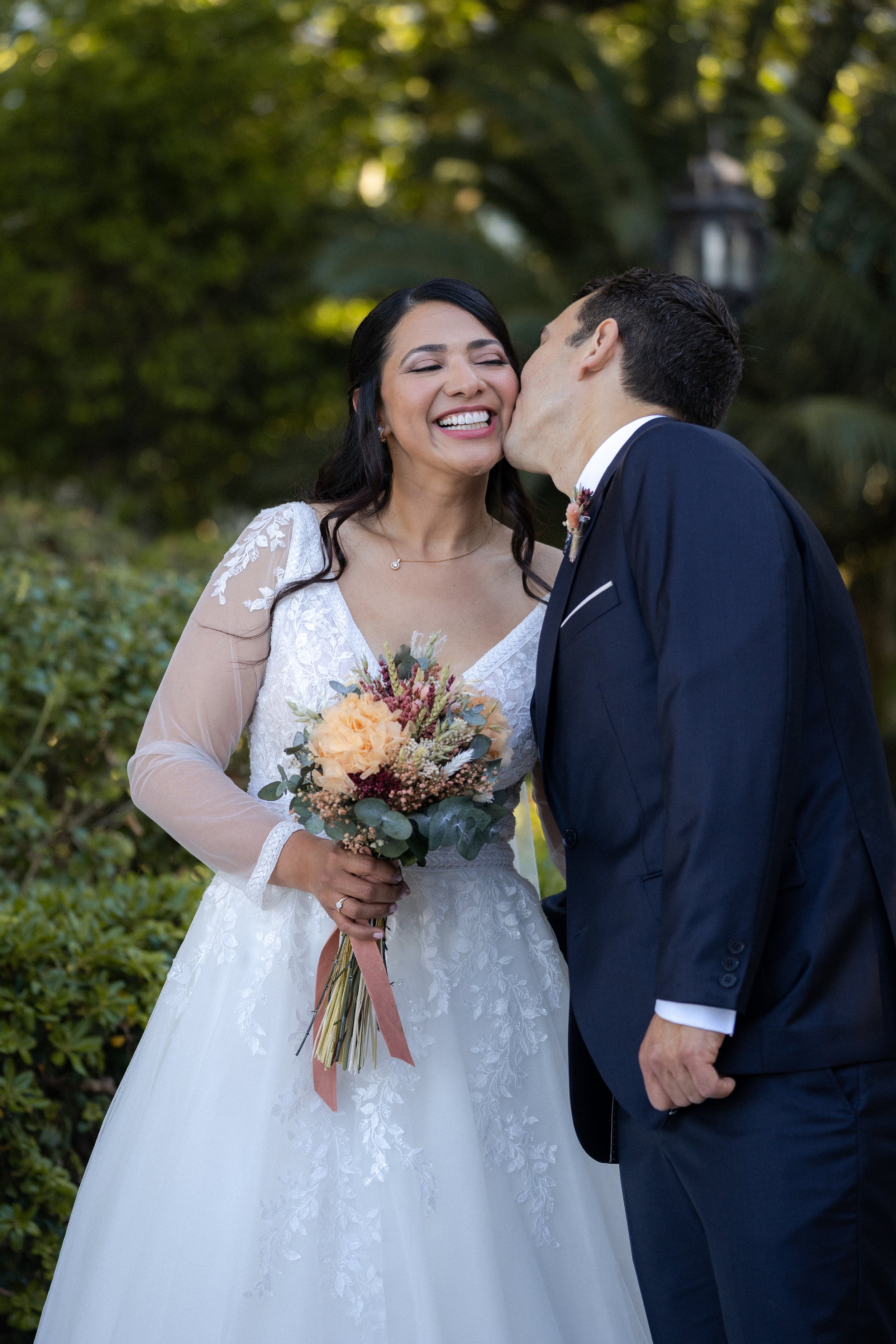 Pareja de recién casados, el hombre besa a la mujer en la mejilla, vestidos de traje y vestido de novia, ella sostiene un ramo de flores.
