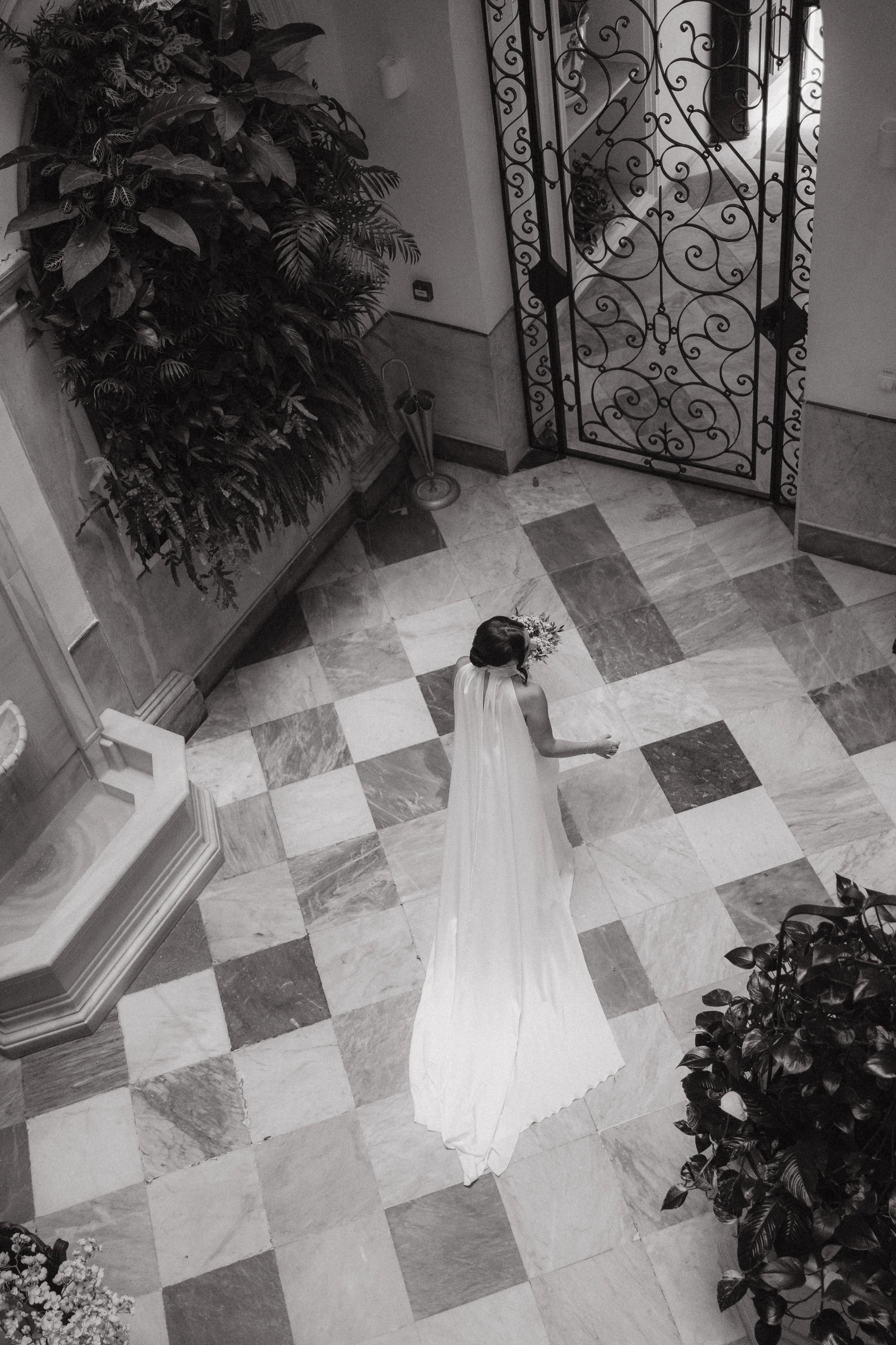 Mujer con vestido de novia en un lugar elegante con piso de baldosas de colores y puertas de hierro decorativas.