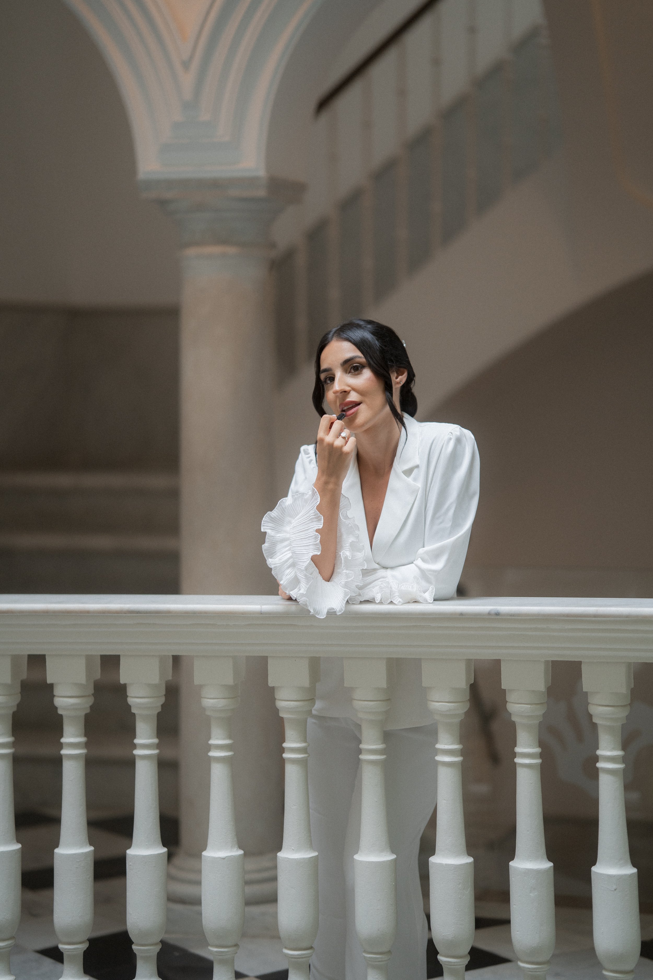 Mujer con cabello oscuro y vestida con blusa blanca, apoyada en una barandilla en un edificio con columnas y arcos.