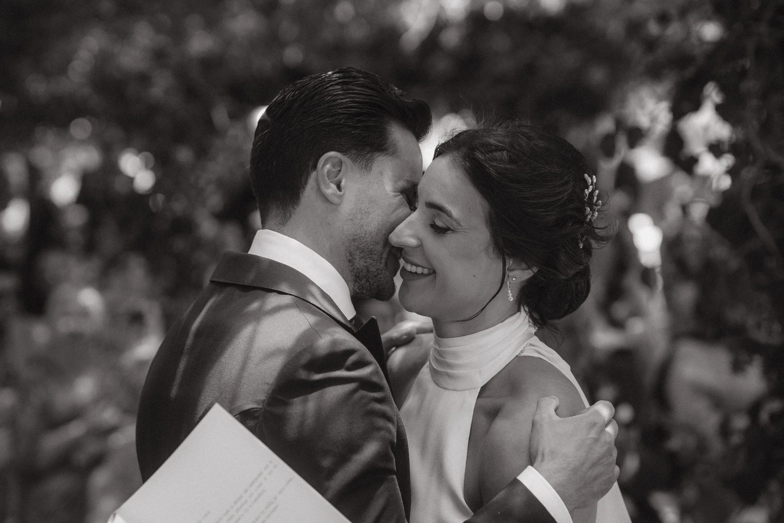 Pareja de novios en una boda, sonriendo y tocándose en un abrazo íntimo, en un entorno natural.
