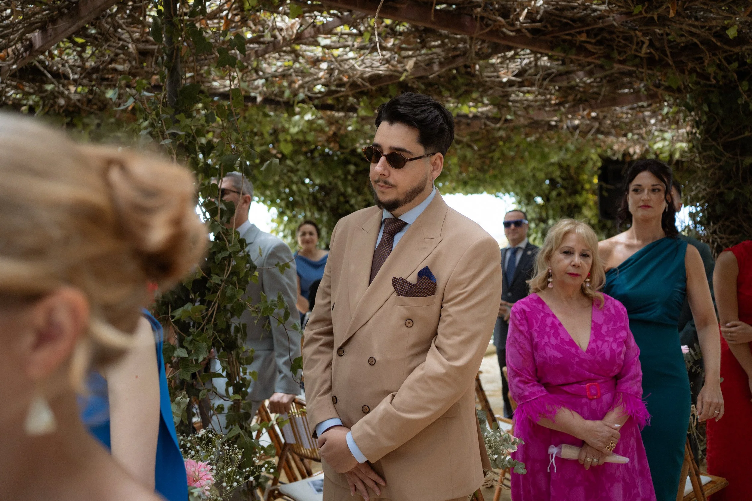 Hombres y mujeres en una ceremonia al aire libre, con algunos arcos decorativos y vegetación en el fondo. Uno de los hombres en primer plano lleva un traje beige, lentes oscuros y está de pie con las manos juntas.