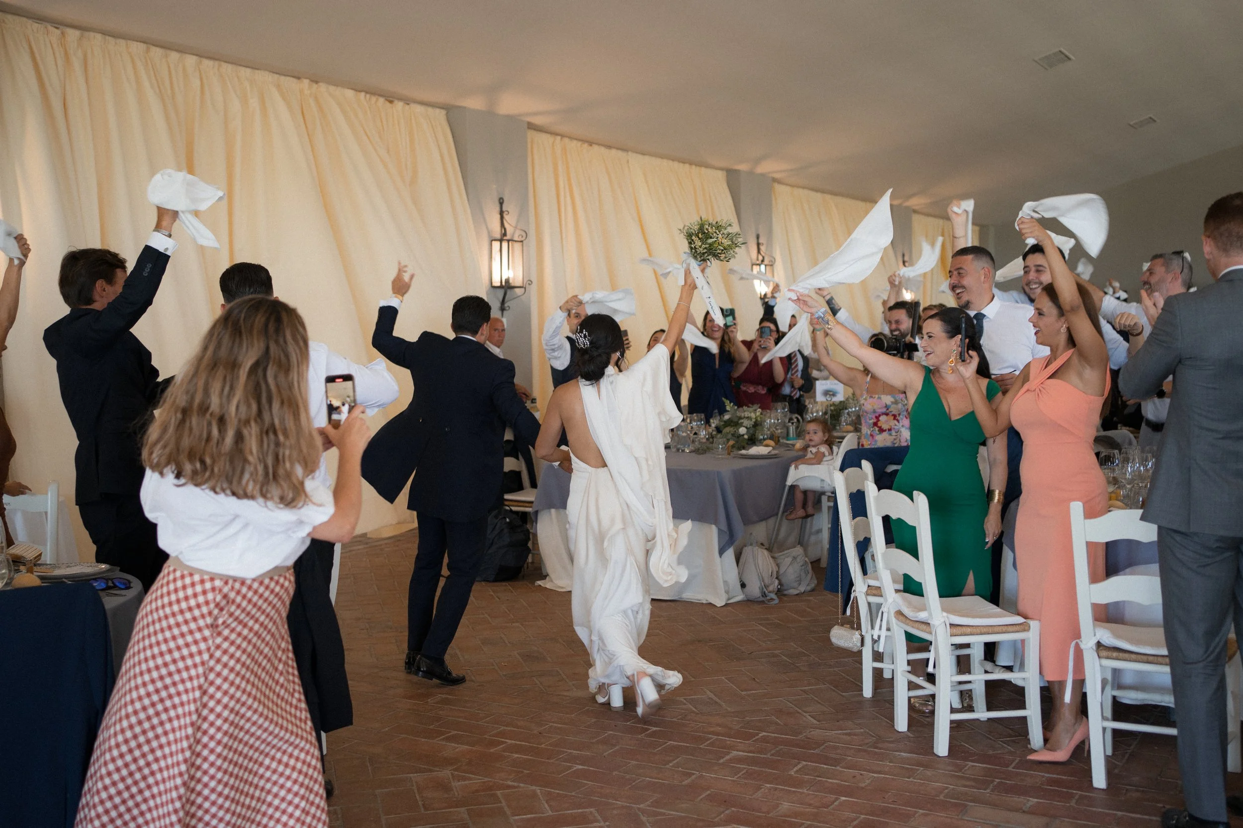 Celebración de boda con invitados lanzando servilletas y felicitando a la novia que porta flores en brazo, en un salón decorado con cortinas y velas.