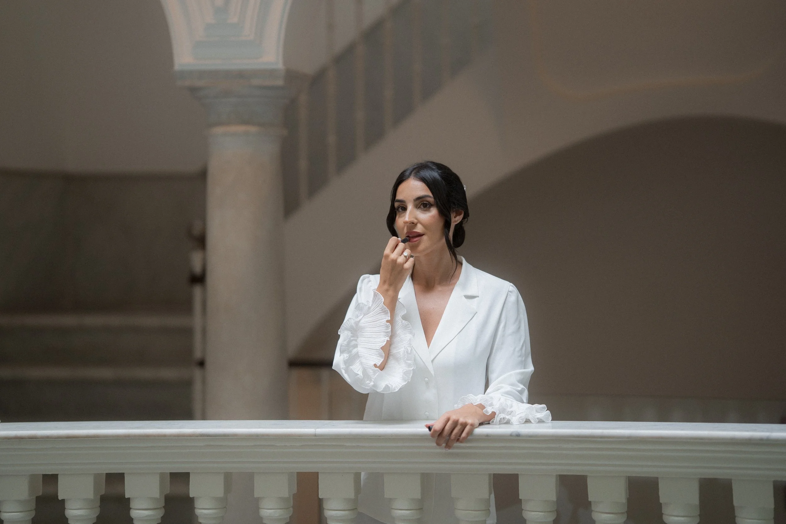 Mujer de cabello oscuro peinada con estilo vintage, vestida con blusa blanca con puños de encaje, en un balcón de un edificio antiguo con columnas de mármol y arcos en la parte superior.