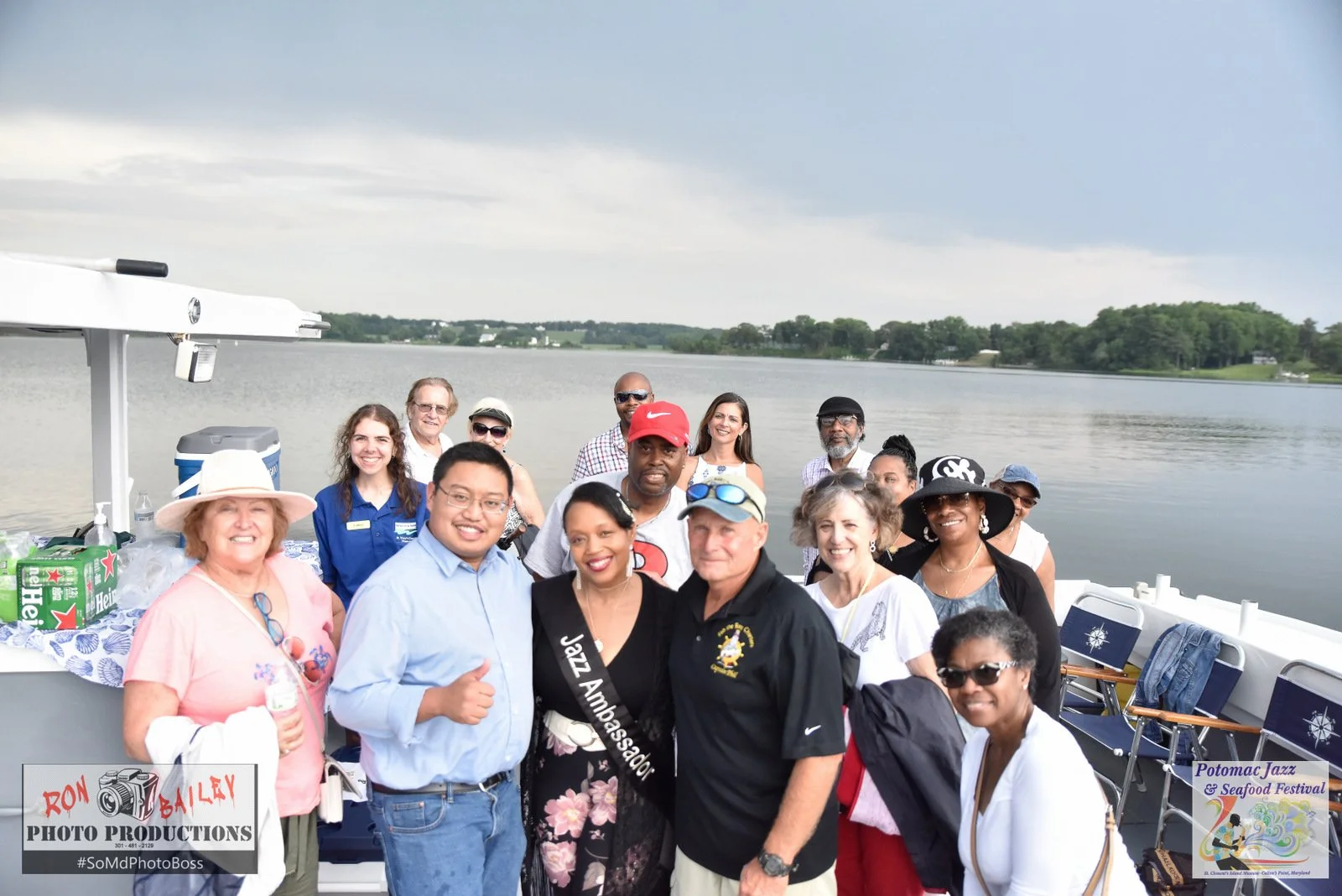 Group of people posing on a boat with a lake in the background, clear sky, and trees lining the far side. Some participants wear festival apparel and a sash reading "Jazz Ambassador." The image includes logos for "Ron Bailey Photo Productions" and "Potomac Jazz & Seafood Festival."