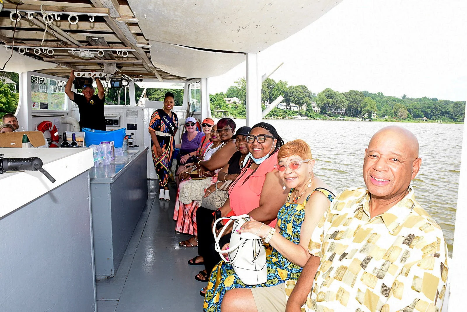 Group of people sitting on a boat smiling, with a person wearing a sash standing nearby, set on a body of water with greenery in the background.