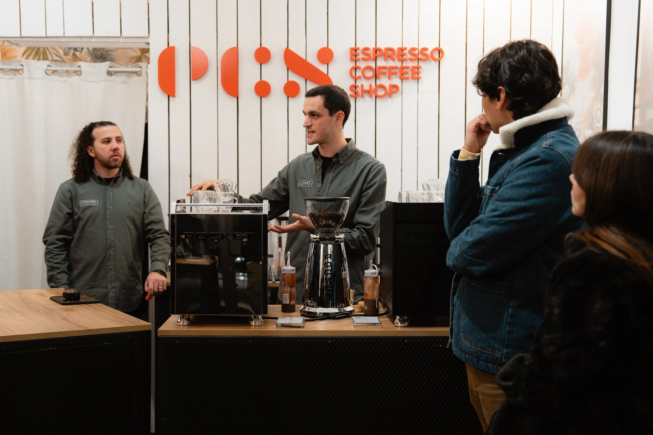 Barista explaining coffee to customers at a coffee shop counter with a sign that reads 'Espresso Coffee Shop' in the background.