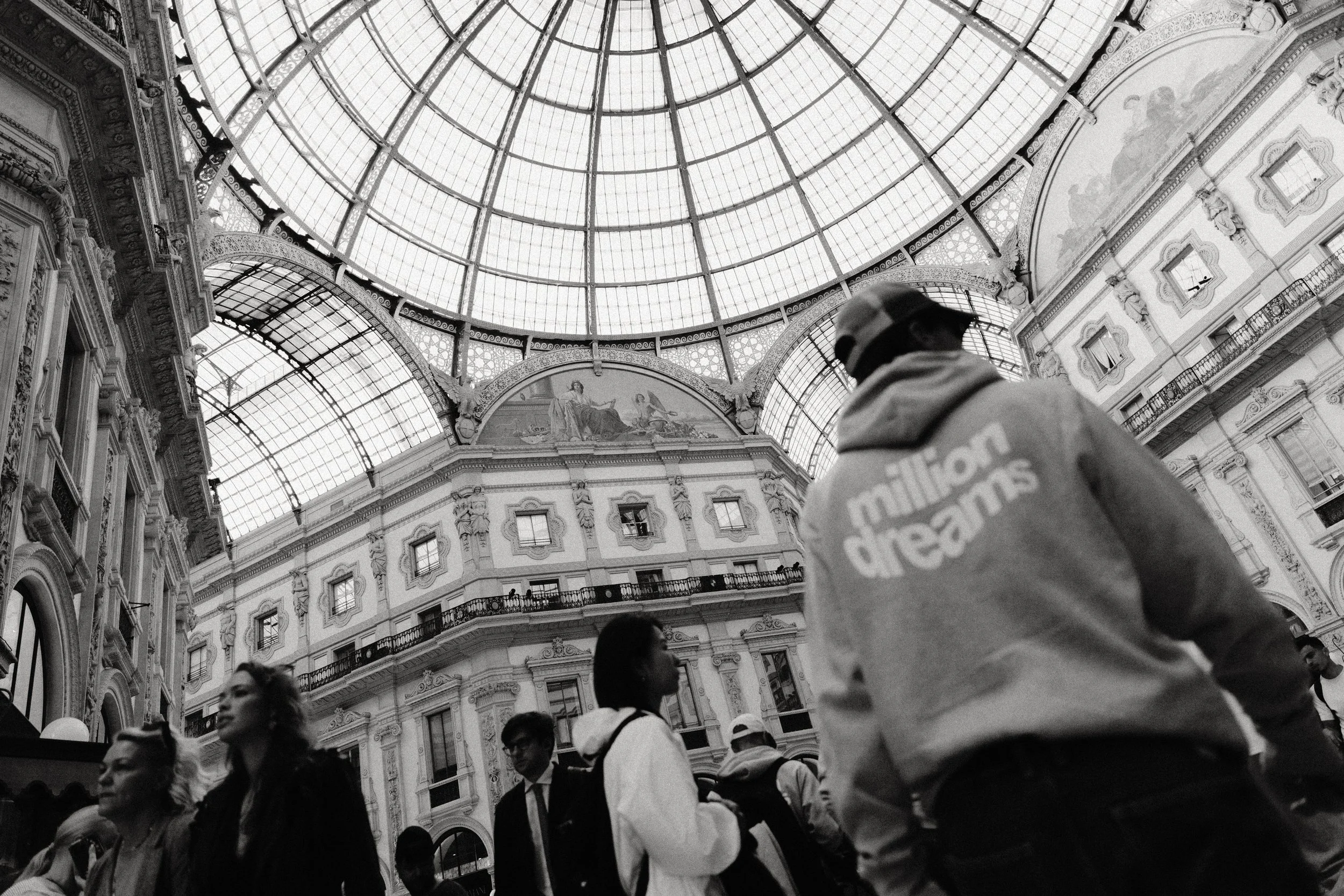 Multiple people walking inside a grand architectural building with a large glass dome ceiling and ornate decor.