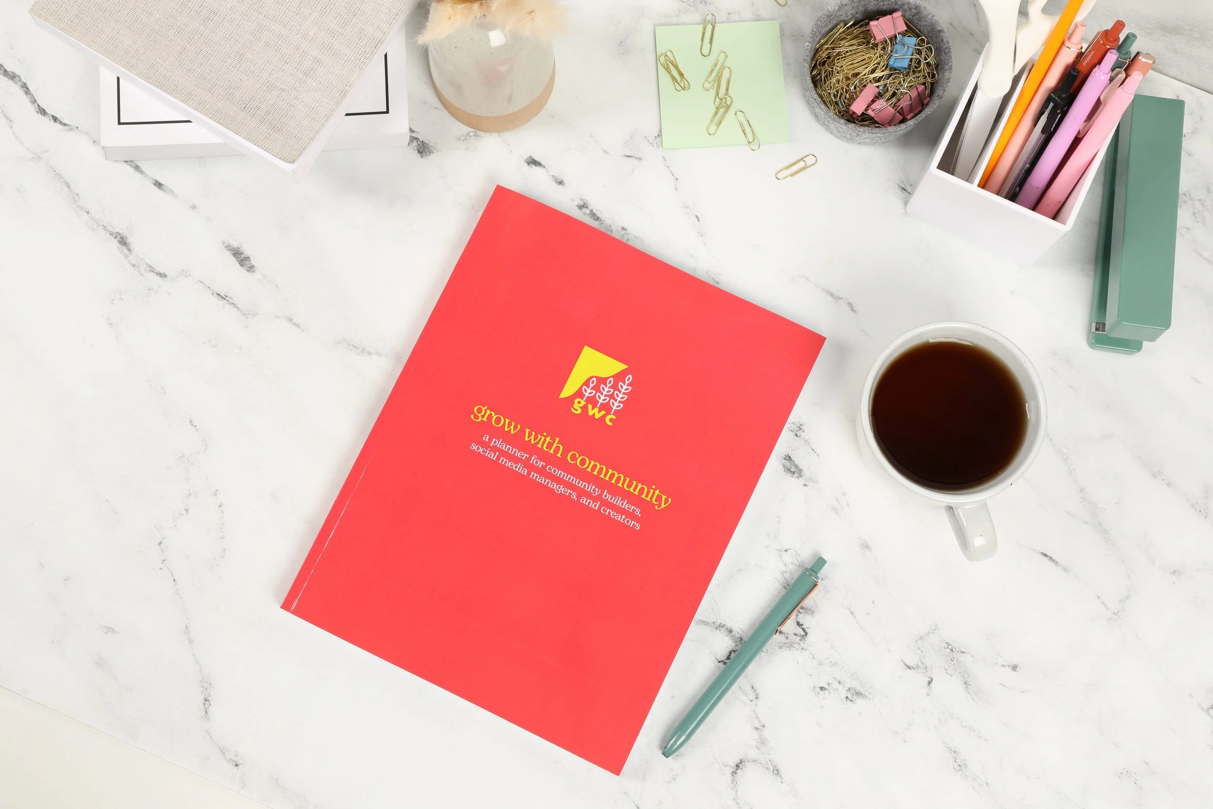 A desk with a red planner titled "Grow with Community" surrounded by office supplies including a cup of coffee, pens, paper clips, sticky notes, and a notebook on a white marble surface. Grow With Community planner sold on Amazon.