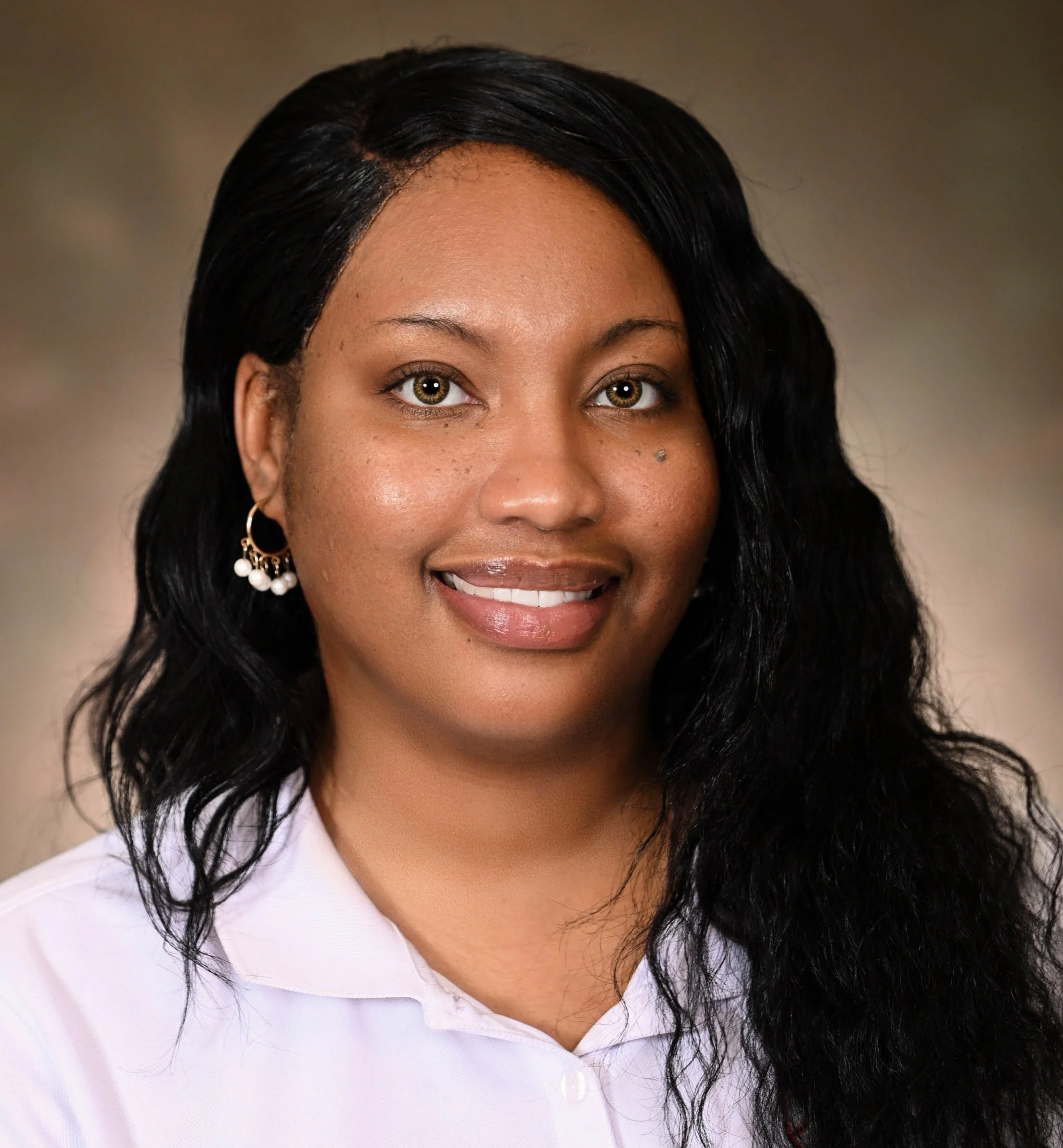 A woman with long black wavy hair, wearing a white shirt and pearl earrings, smiling in a professional portrait.