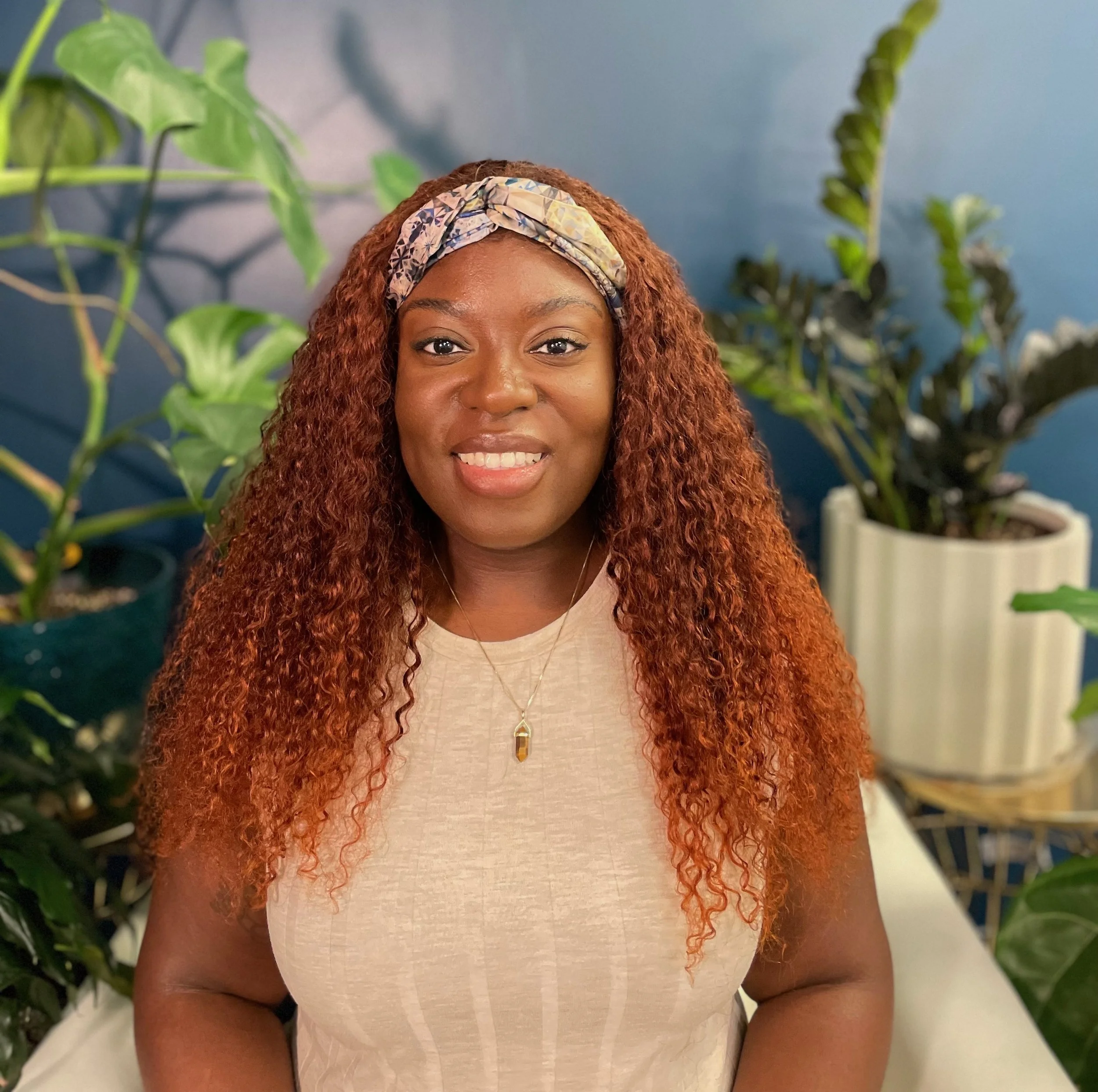 A smiling African American woman with curly, reddish-brown hair wears a beige top, a colorful headband, and a gold necklace with a pendant, sitting among green houseplants against a blue wall.