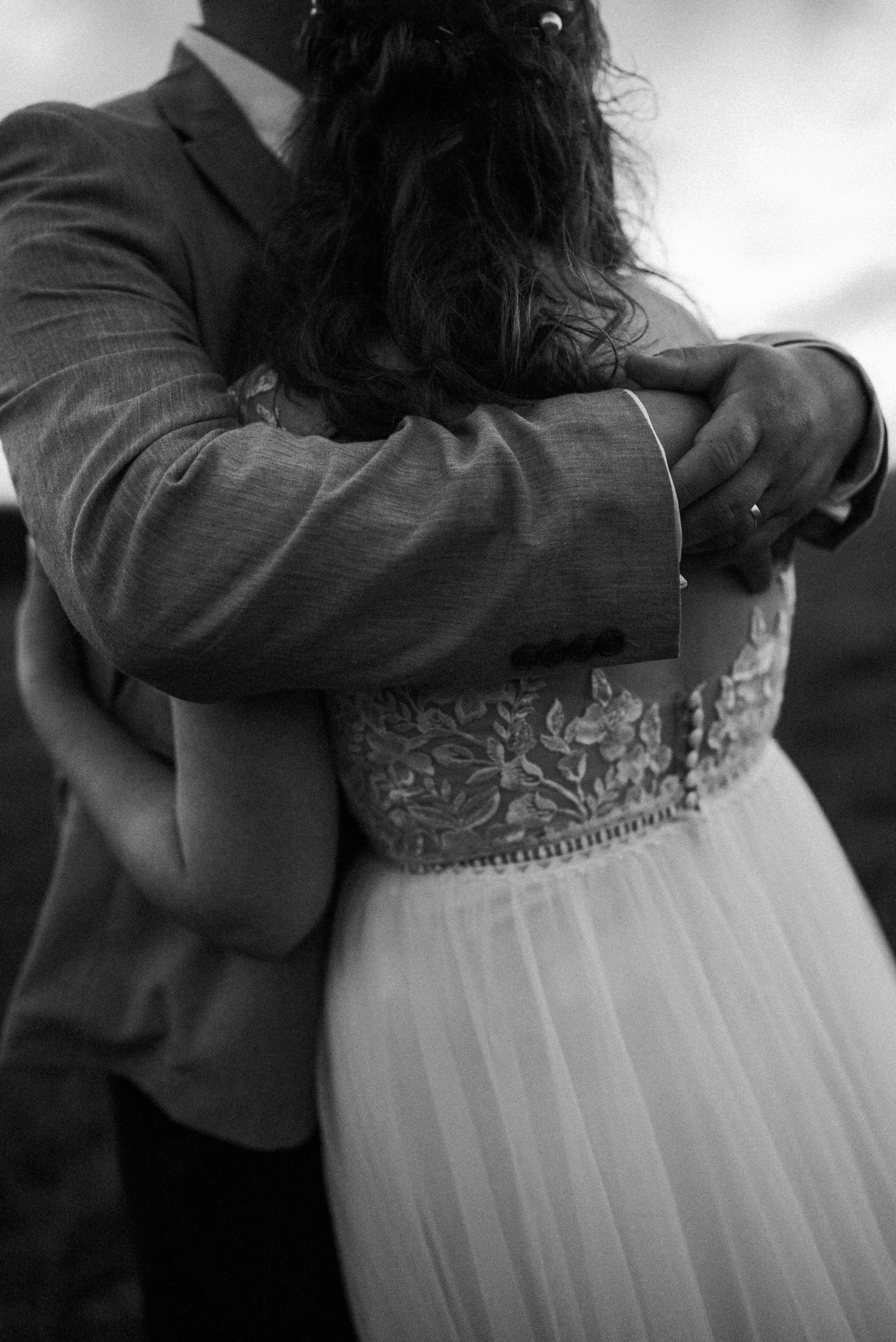 Black and white photo of a couple embracing, showing a close-up of their torsos. The woman wears a detailed, embroidered dress and the man is in a suit.