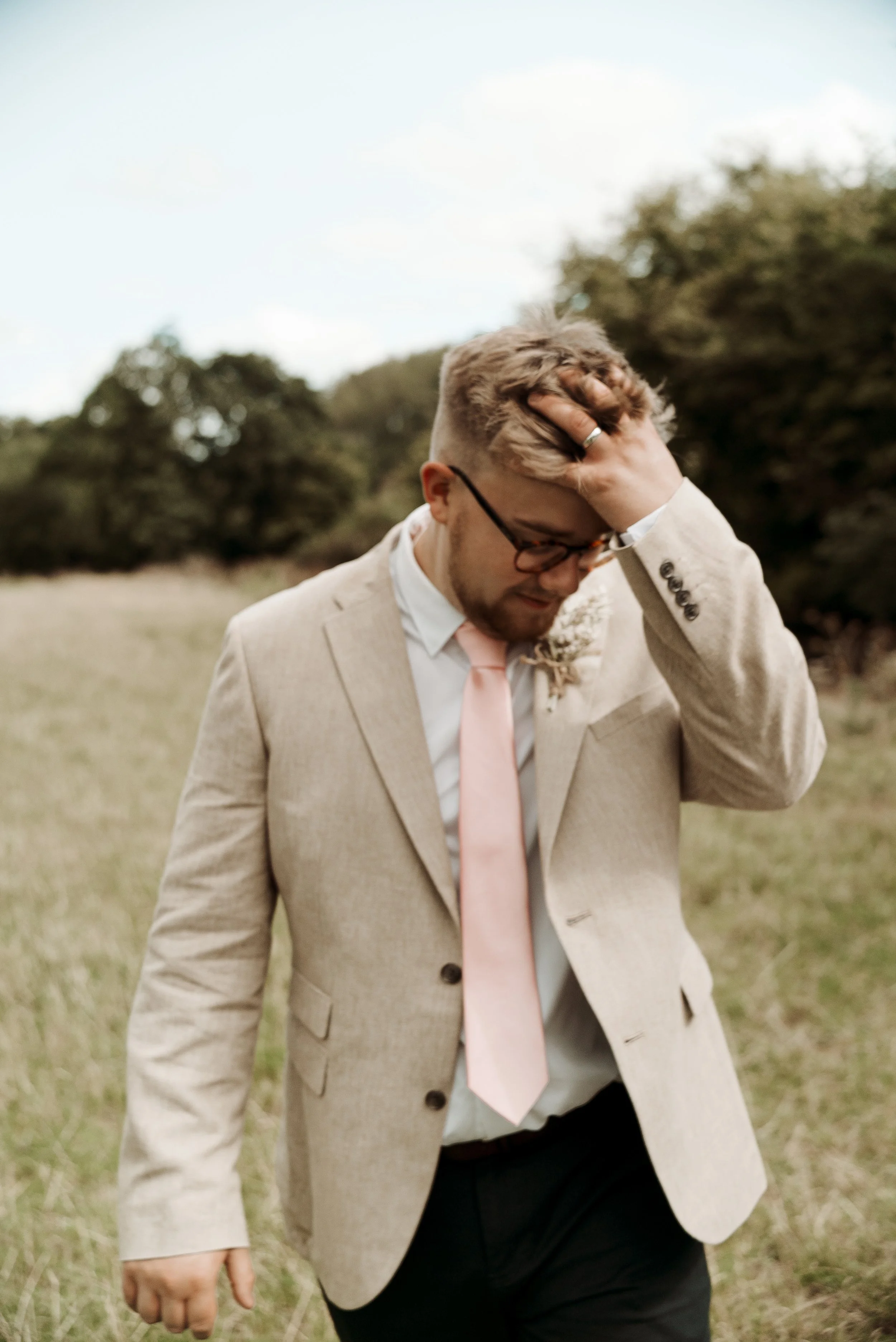 Man in beige suit with pink tie standing in grassy field with trees in background, hand in hair, wearing glasses.