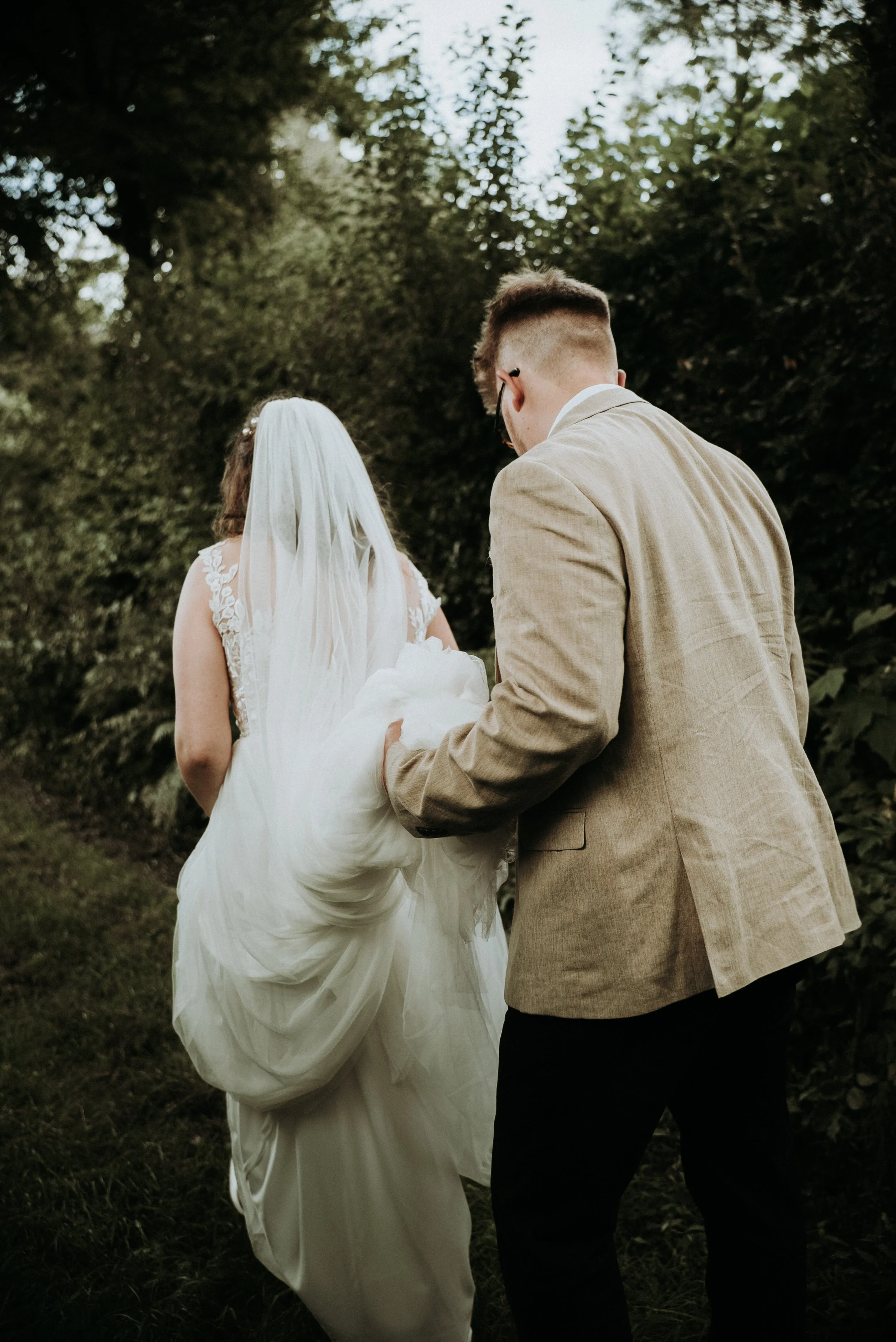 Bride and groom walking outside, groom holding bride's dress, surrounded by greenery.