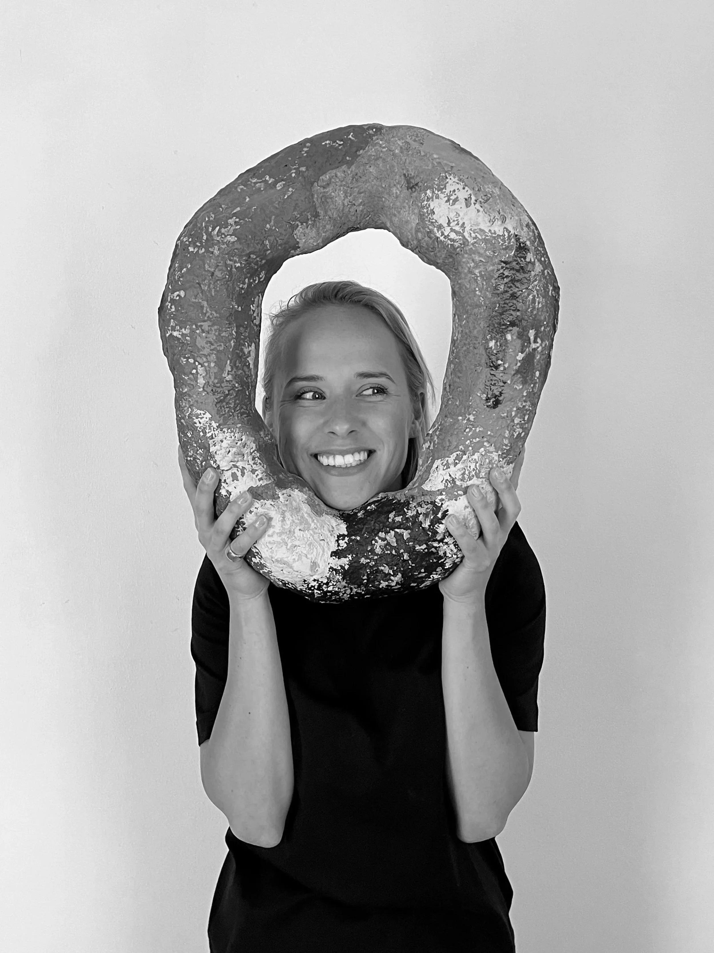 A woman smiling and holding a large, round, baked bread or pastry shaped like a doughnut around her face.