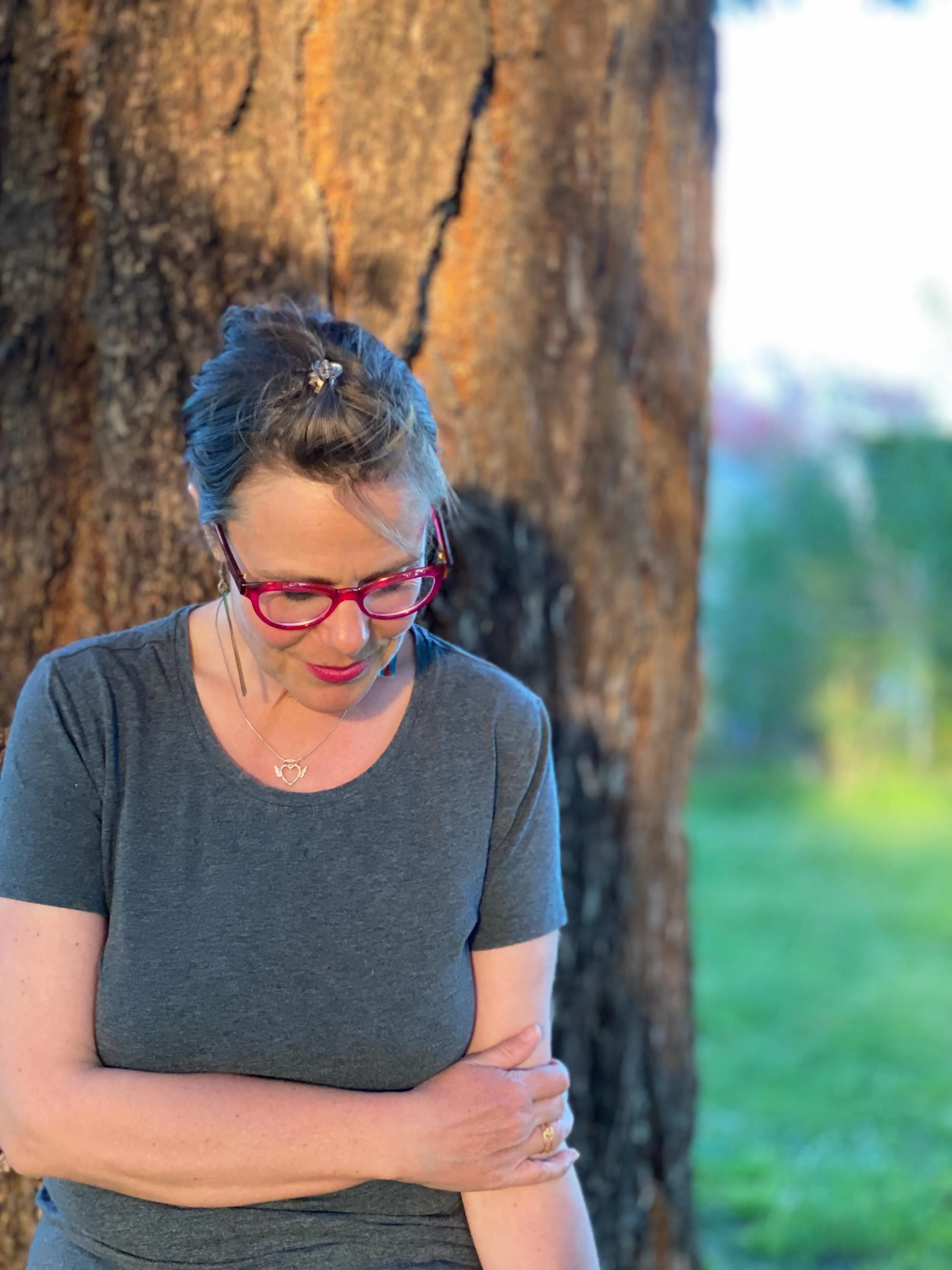 Ria de Neeve, astrologer, in front of a tree wearing grey t-shirt looking down