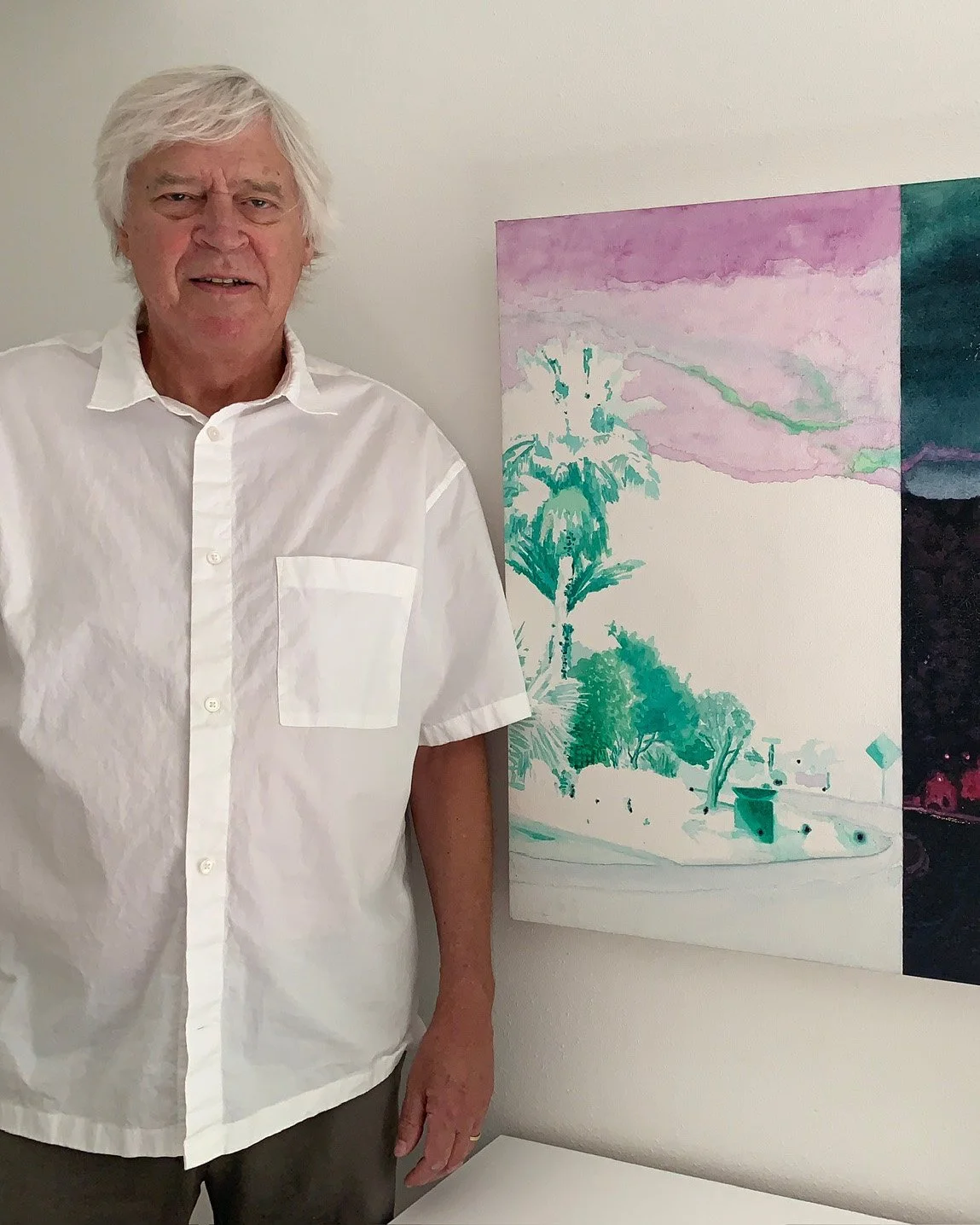A man with gray hair in a white short-sleeve button-up shirt standing next to a colorful abstract landscape painting on the wall.
