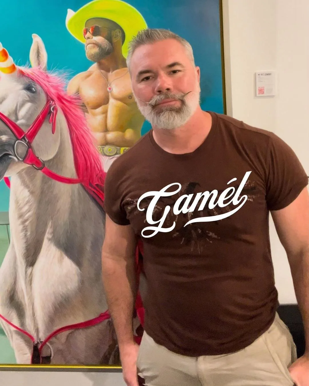 A man with gray hair and beard standing in front of a colorful painting of a muscular man with sunglasses and a yellow hat, and a white horse with pink mane and harness.