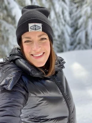 A woman in a black beanie and puffer jacket smiling in a snowy forest, representing tips on taking a good selfie.