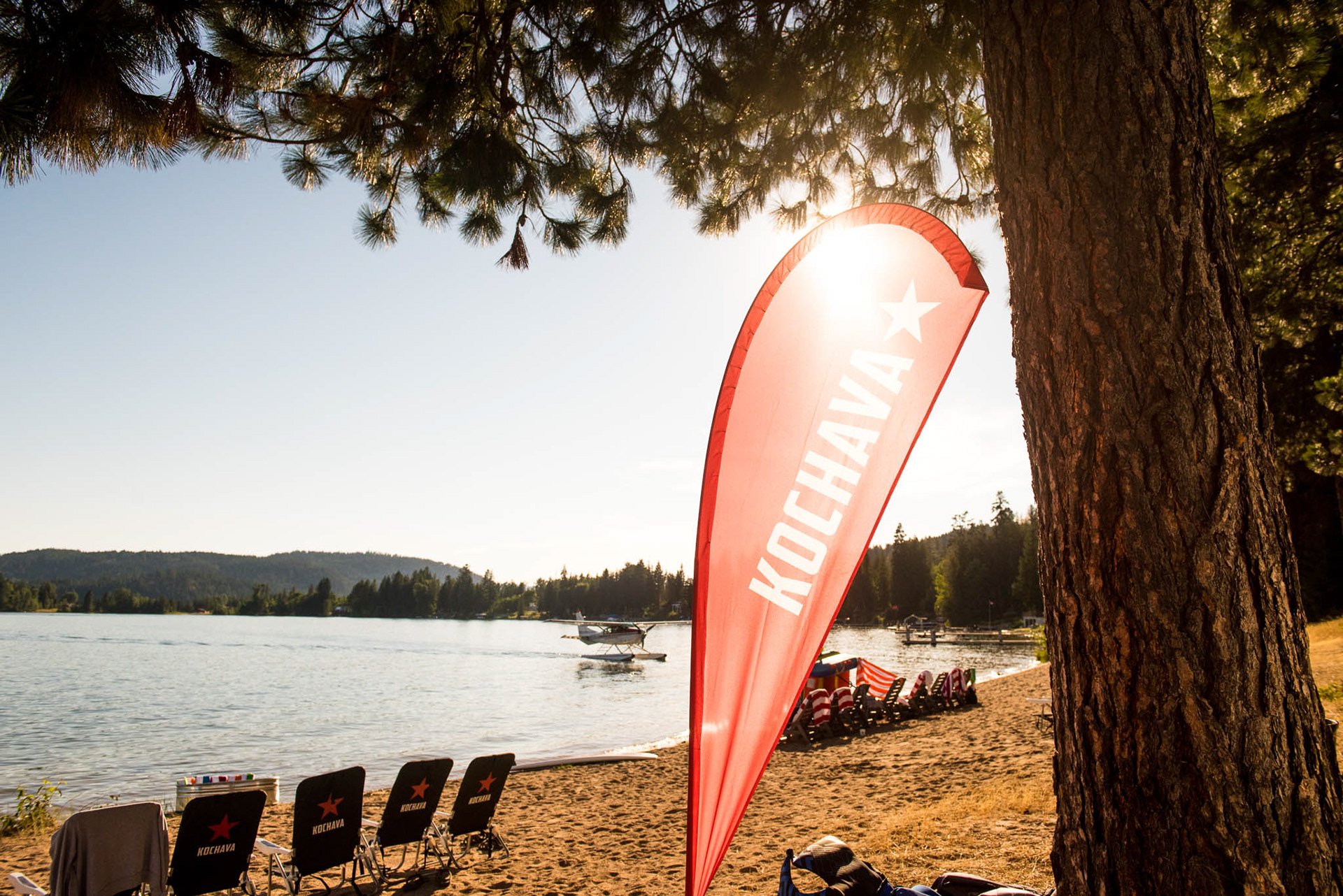 A lakeside scene with a red Koshava flag, beach chairs, and boats on the water, under the sun and trees.