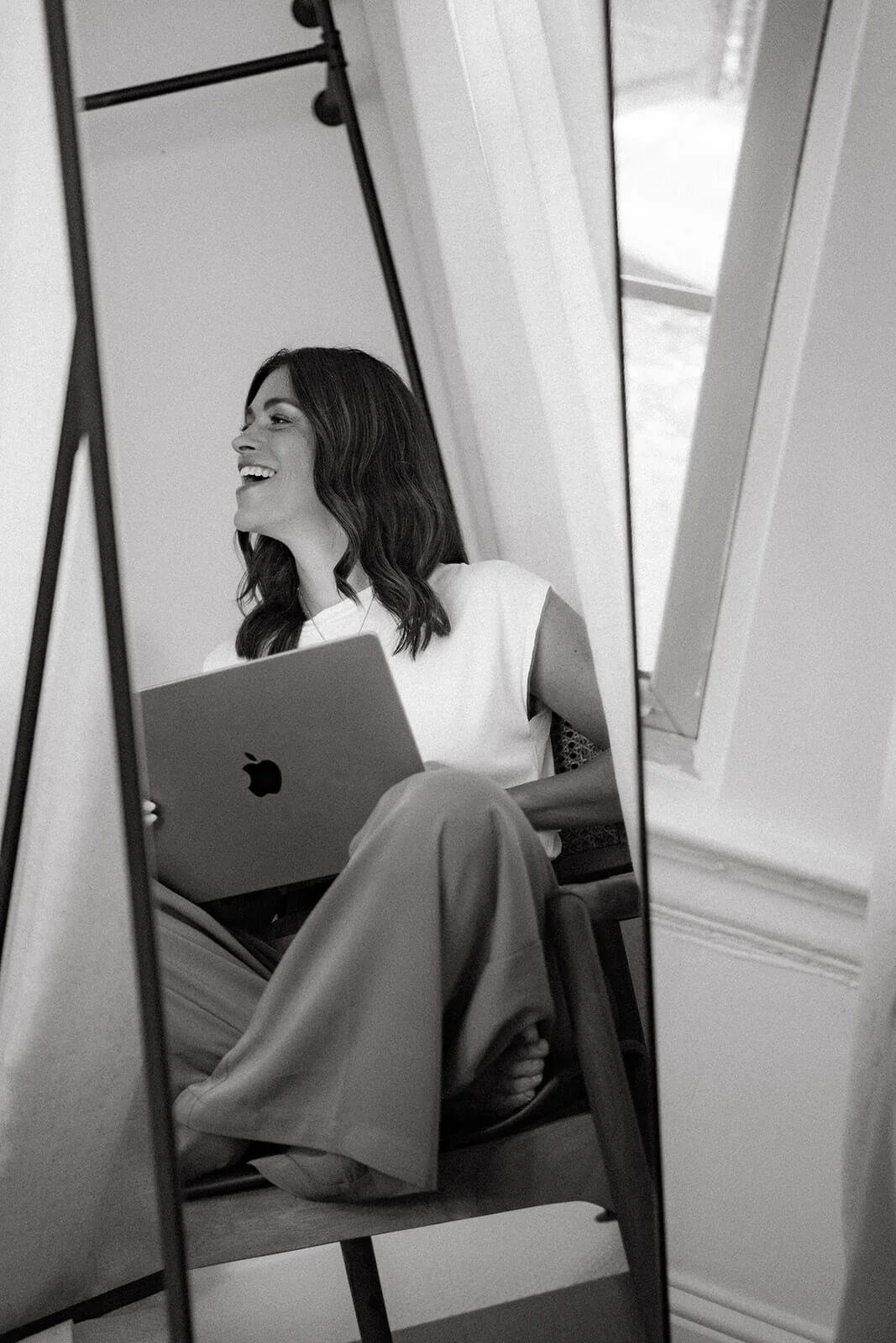 Jane Gion sitting in a chair, smiling and looking to her left, holding a laptop, reflected in a mirror.