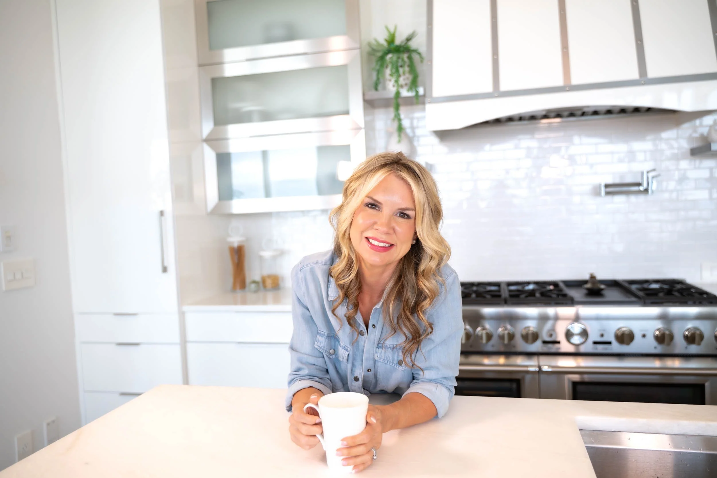 A woman with long wavy blonde hair smiling and holding a white mug in a modern white kitchen.