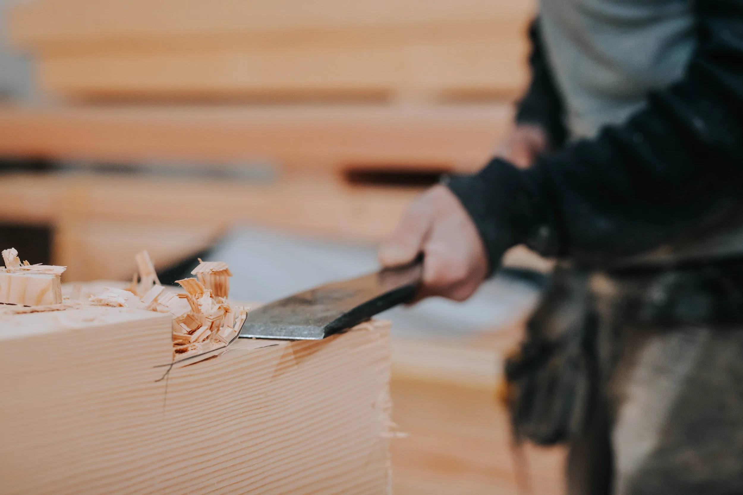 close-up of a person's hands using a chisel to carve and shape a piece of wood, creating wood shavings.
