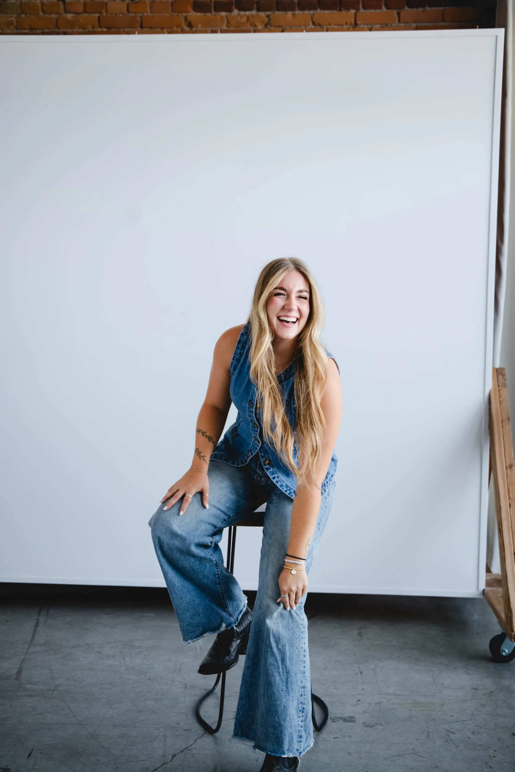 Emilie Tangeman with long blonde hair, smiling and laughing, sitting on a black stool in front of a white backdrop, wearing a sleeveless denim vest, wide-leg jeans, and black boots, with a brick wall and wooden ladder in the background.