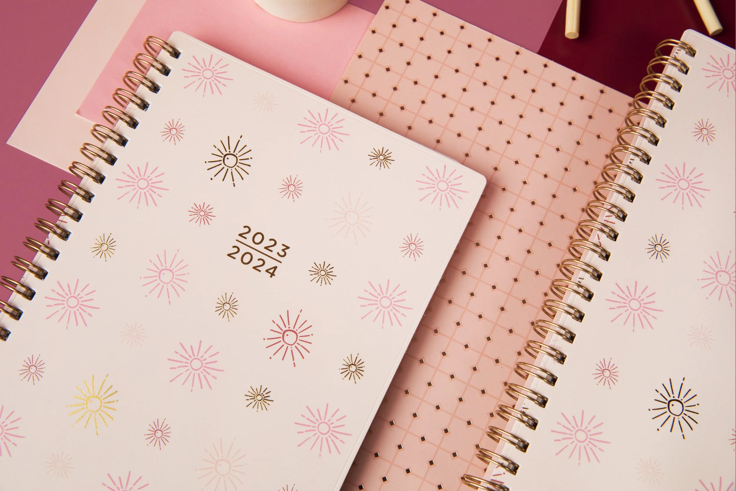Close-up of pink and white spiral-bound planners with firework patterns, stacked on a pink and burgundy background.