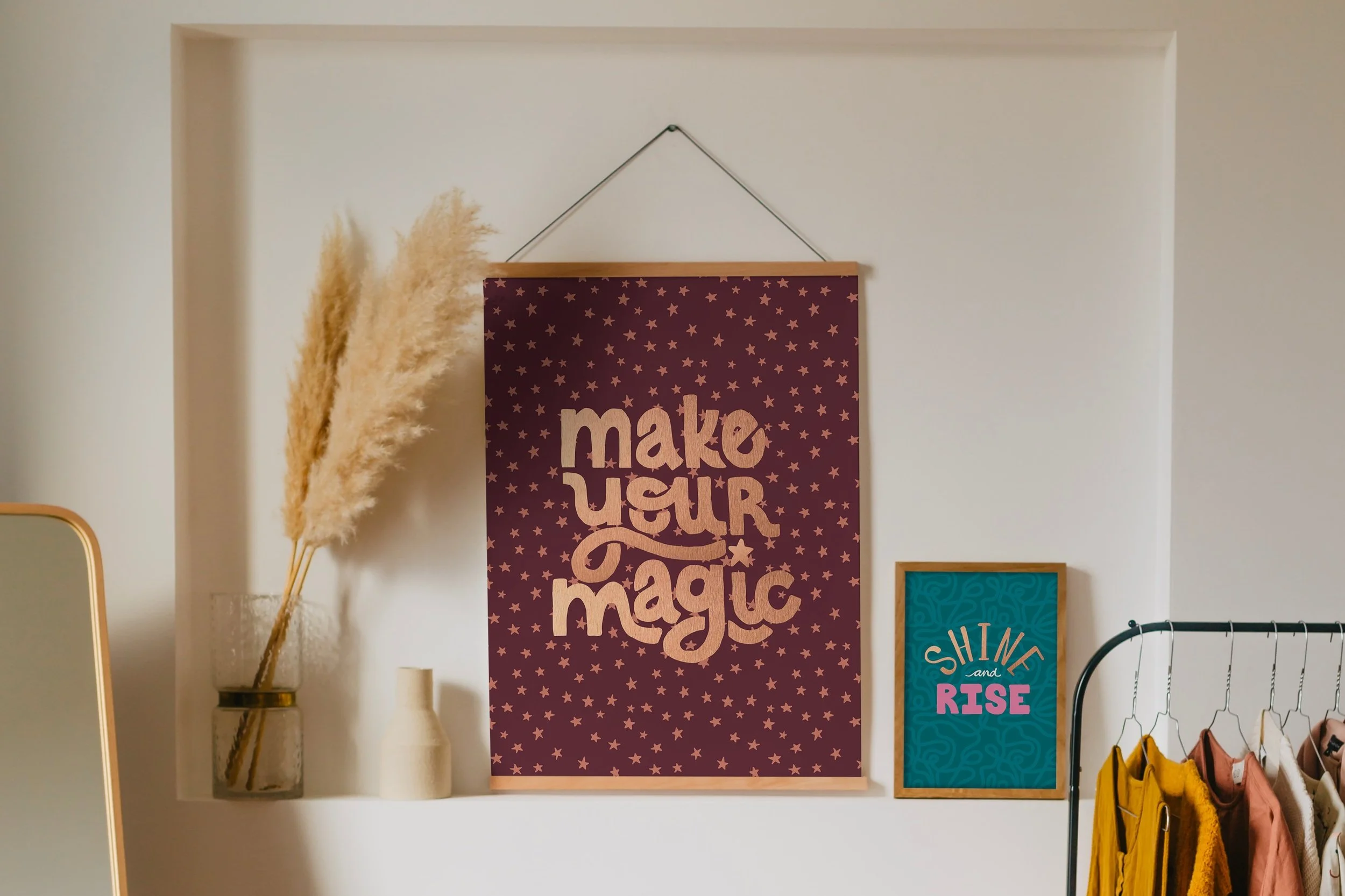Decorative wall display featuring a large burgundy poster with gold lettering and star patterns, flanked by two framed posters, and a tall glass vase with pampas grass on a shelf.