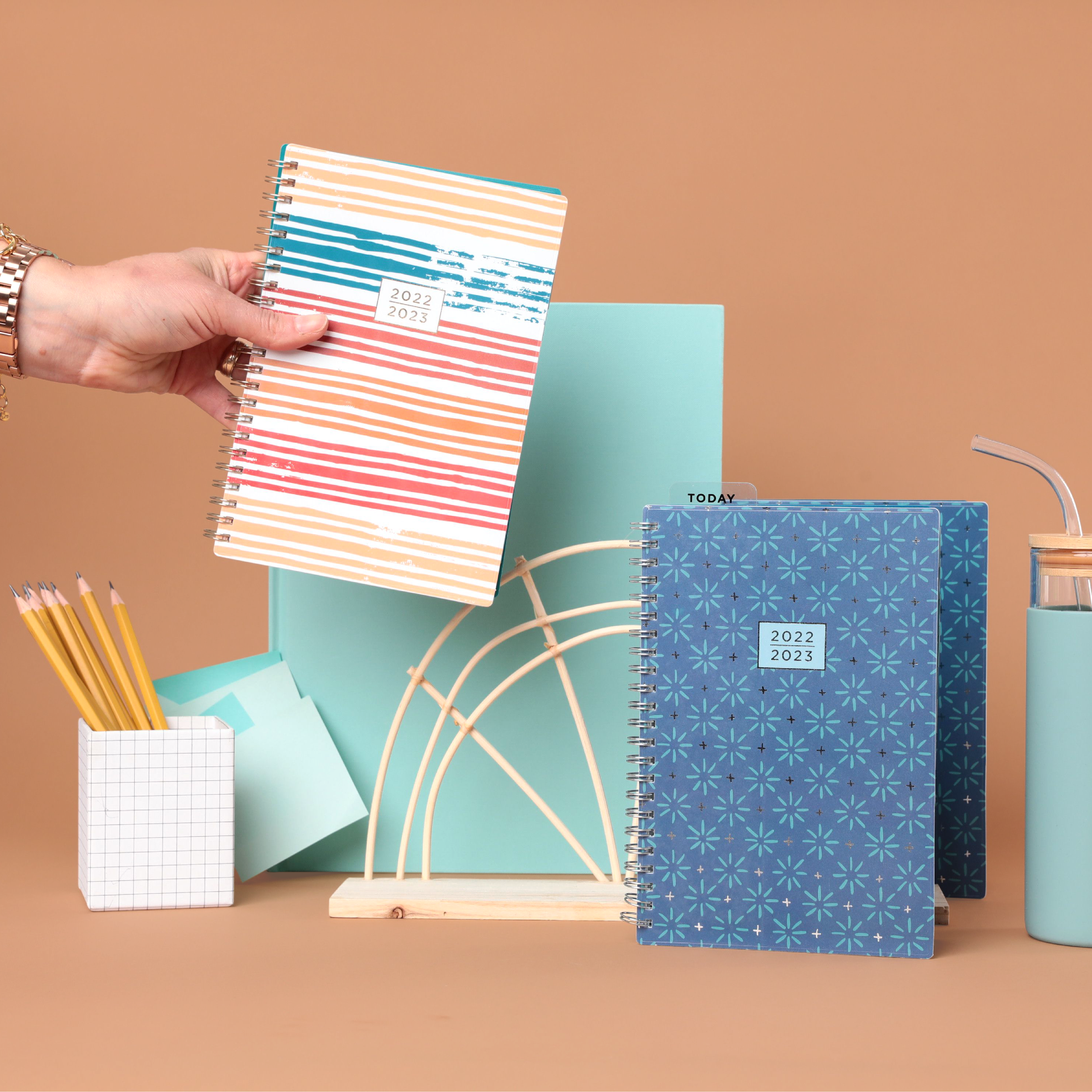 A collection of notebooks, planters, pencils, and a glass container arranged on a pastel-colored surface. The notebooks have colorful covers, some with patterns and labels indicating academic years, and are placed with some standing upright and other
