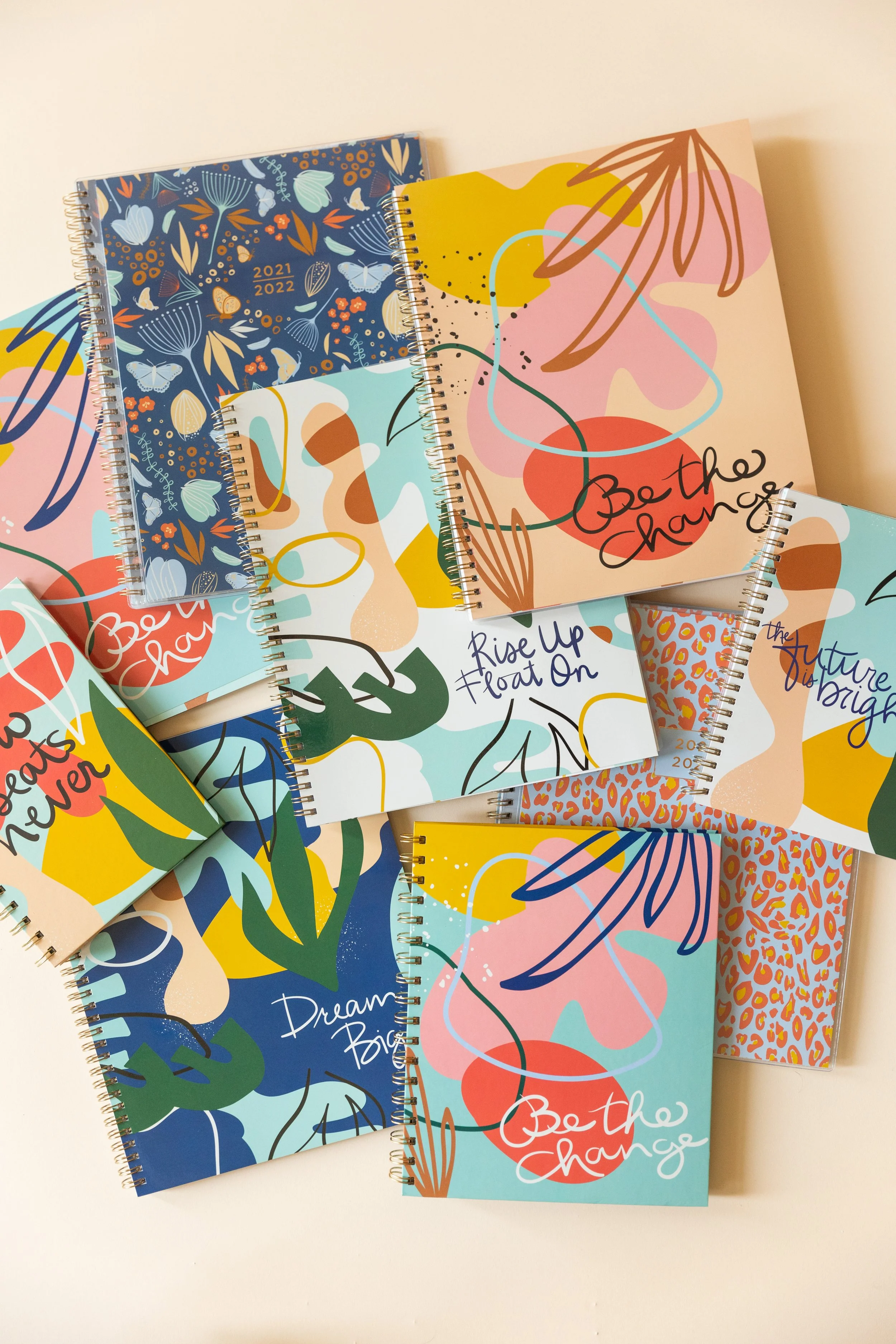 A collection of colorful spiral-bound notebooks with abstract and floral designs, some with inspirational quotes like "Be the change" and "Dream big," arranged on a white surface.