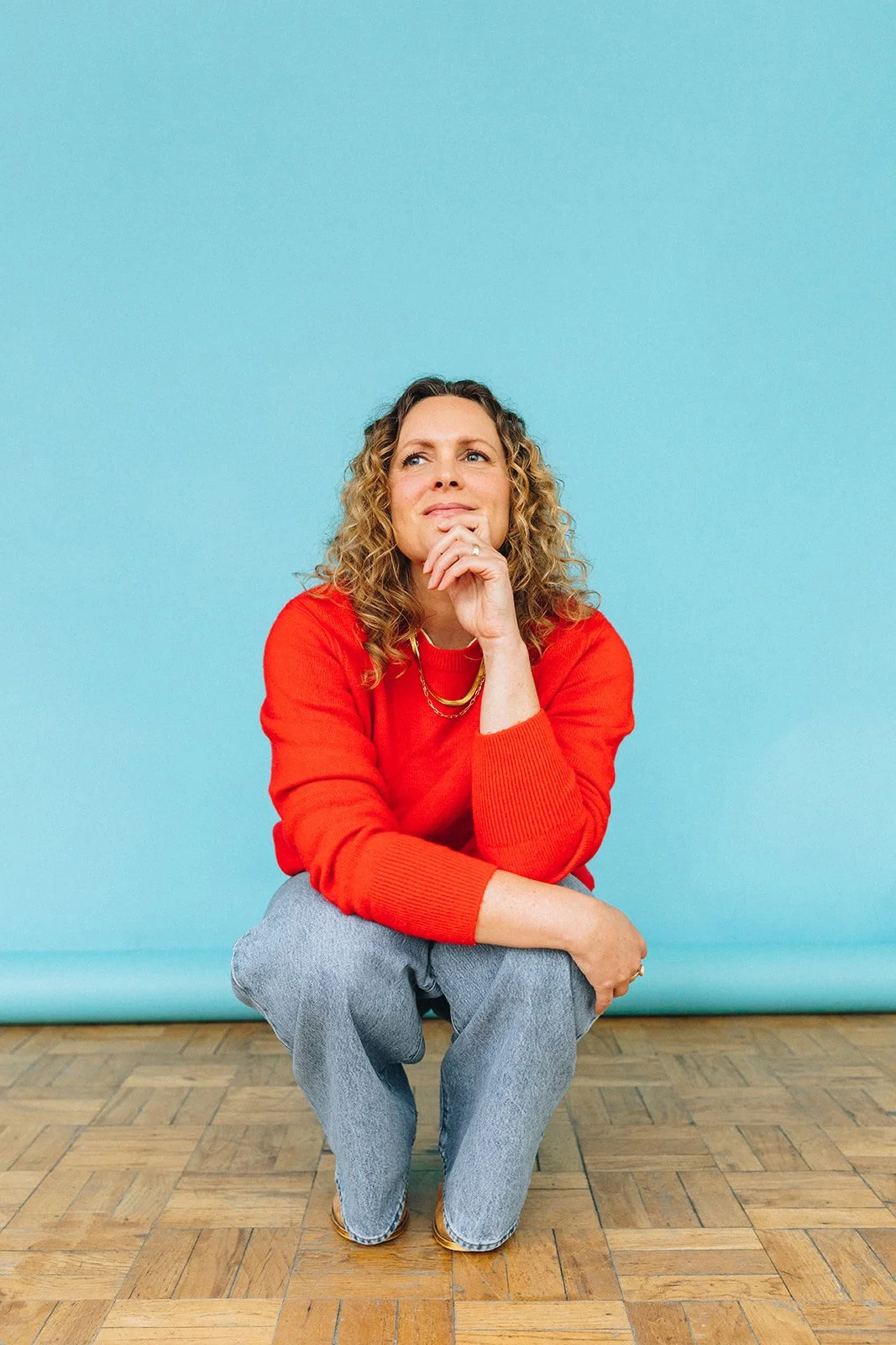 A woman with curly hair wearing a red sweater and gray pants, squatting in front of a light blue background, looking pensively to the side with her chin resting on her hand.