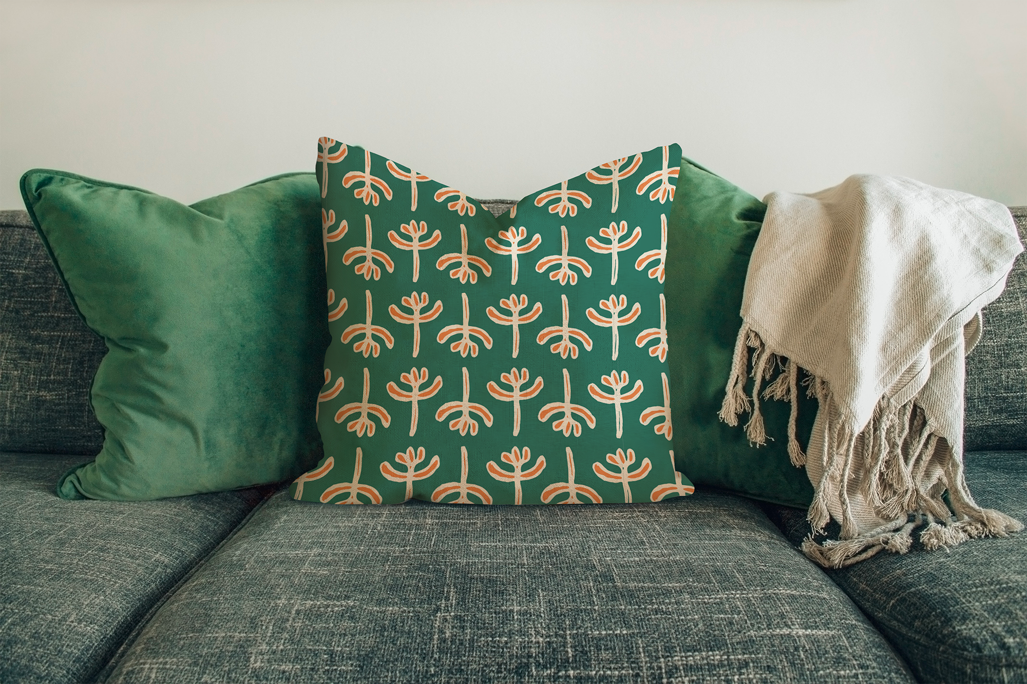 A sofa decorated with three cushions: one solid green cushion on the left, a patterned cushion with a green background and an orange and white abstract design in the middle, and a cream-colored textured blanket on the right.