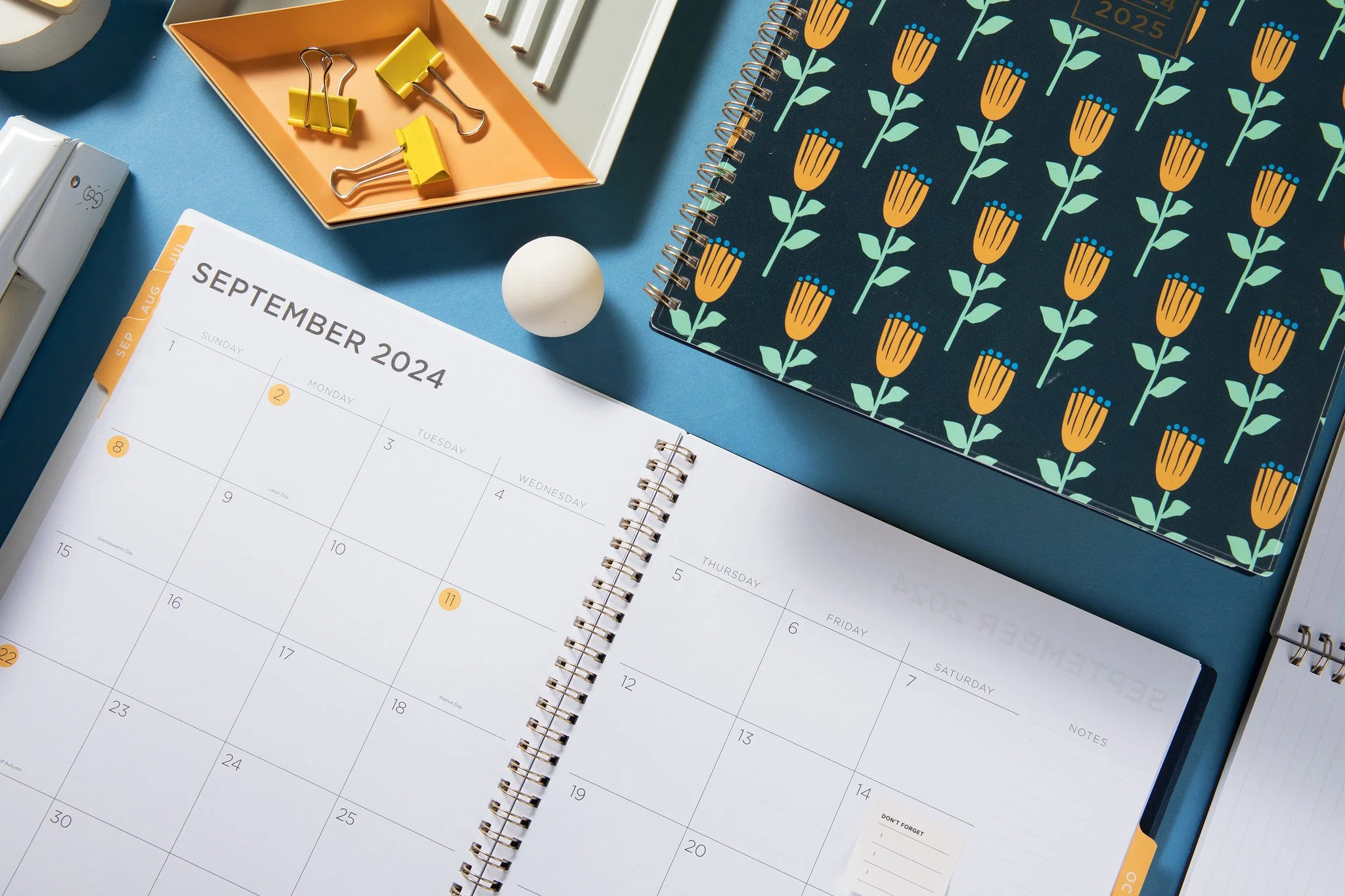 Open planner showing September 2024 calendar with other stationery items like a notepad, binder clips, and a tape dispenser on a blue surface.