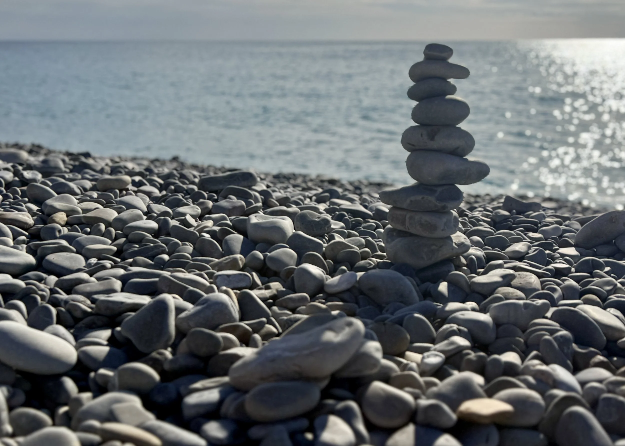 Stacked gray stones on a pebble beach with water and sunlight in the background.