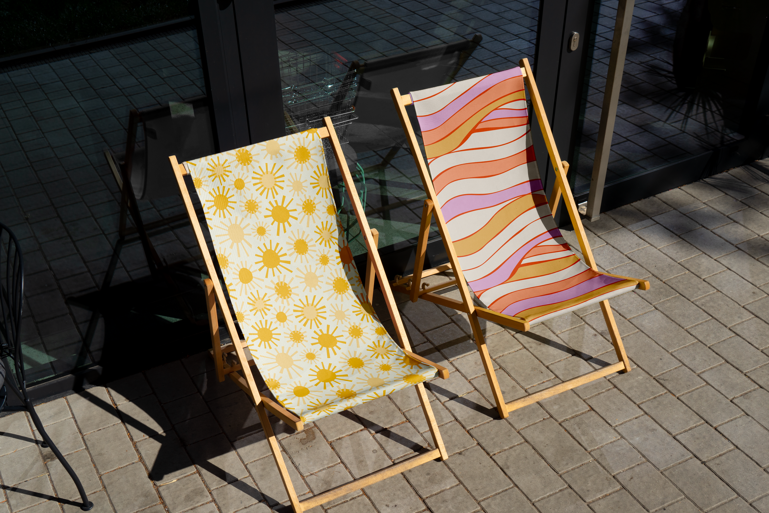 Two wooden deck chairs with colorful striped and yellow floral patterned fabric cushions are positioned on a brick patio outside a glass door.