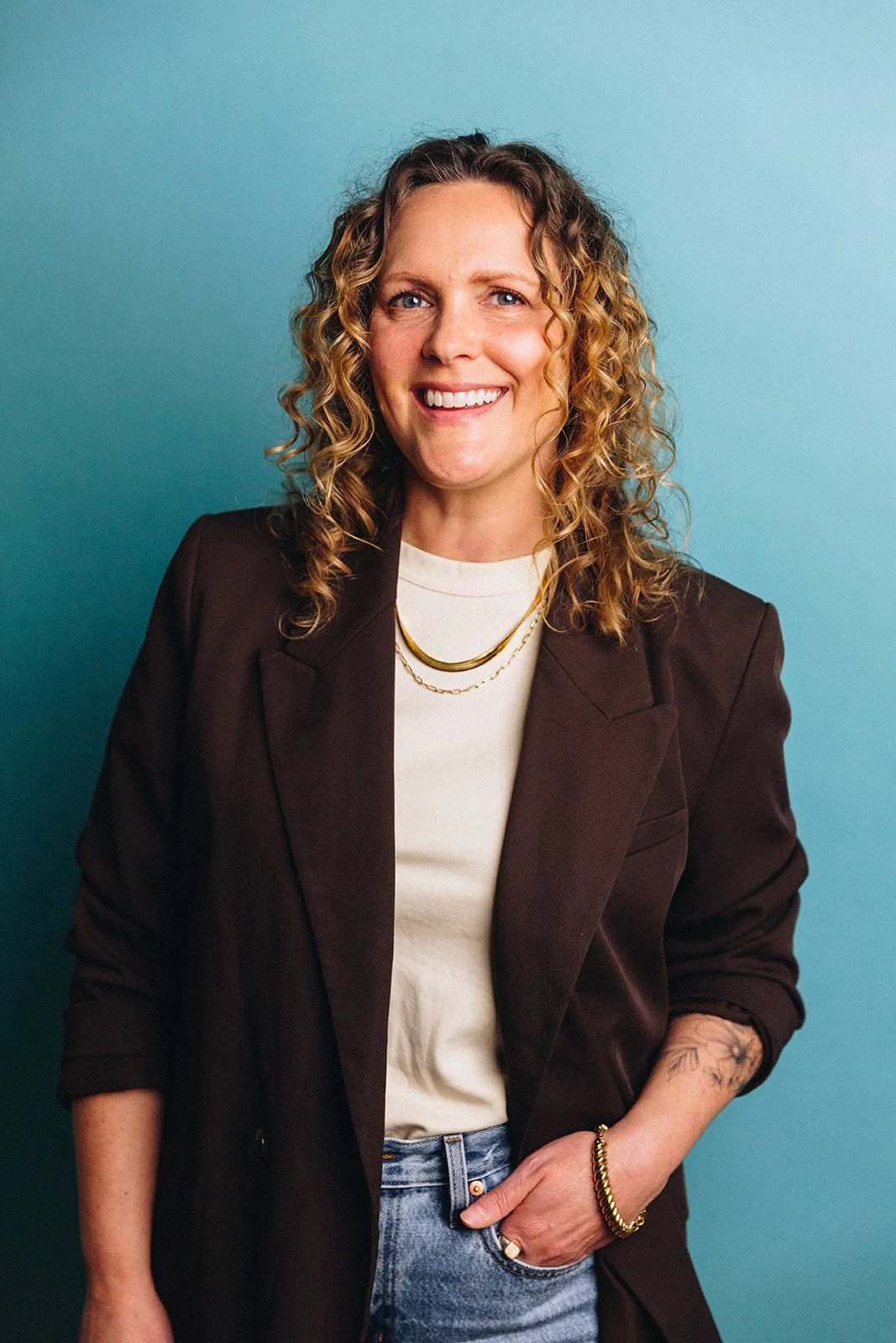 A woman with curly blonde hair smiling, wearing a white t-shirt, brown blazer, and jeans, with layered gold necklaces, a bracelet, and a tattoo on her right forearm, standing against a blue background.