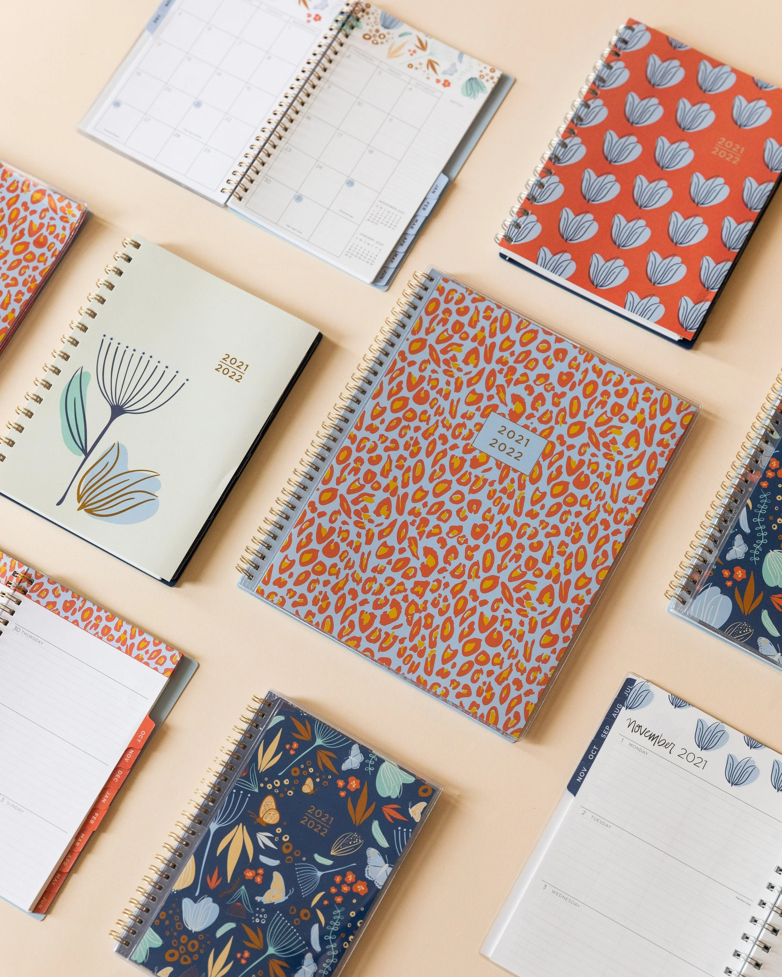 Several 2021-2022 planners with colorful floral and animal print covers are open on a beige surface. Some pages show monthly calendars with blank spaces for dates and notes, and others have weekly or daily planning layouts.