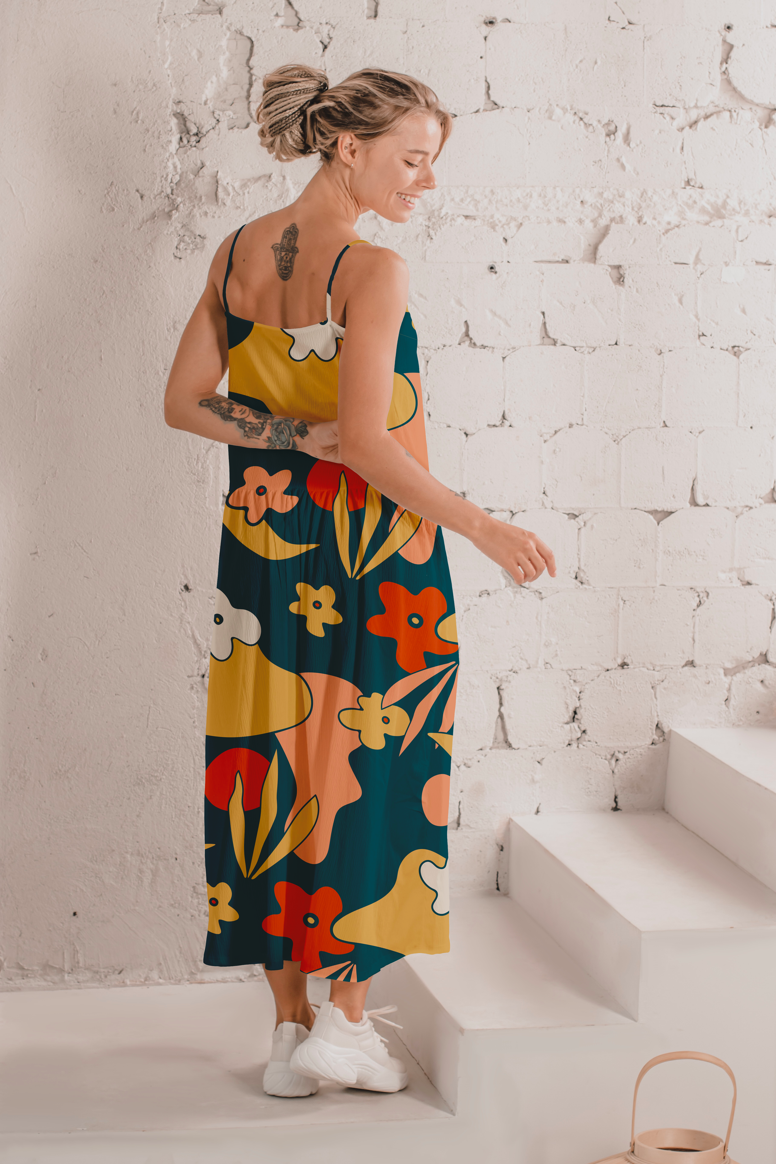 Young woman with dreadlocks wearing a colorful, patterned maxi dress and white sneakers, standing near white stairs against a white brick wall, smiling and looking down.