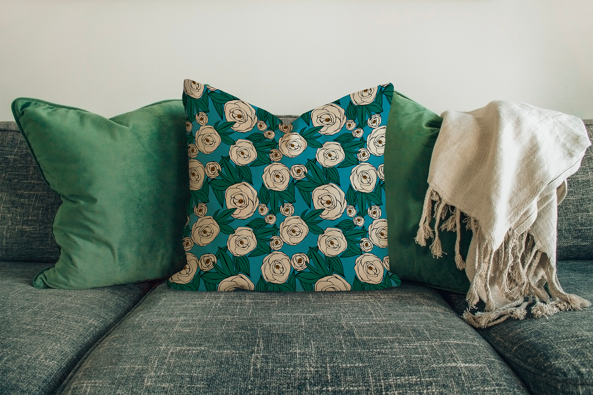 A sofa with two solid green pillows, a floral patterned pillow with cream-colored flowers on a teal background, and a cream-colored throw blanket with fringes draped over the arm of the sofa. The background is plain.