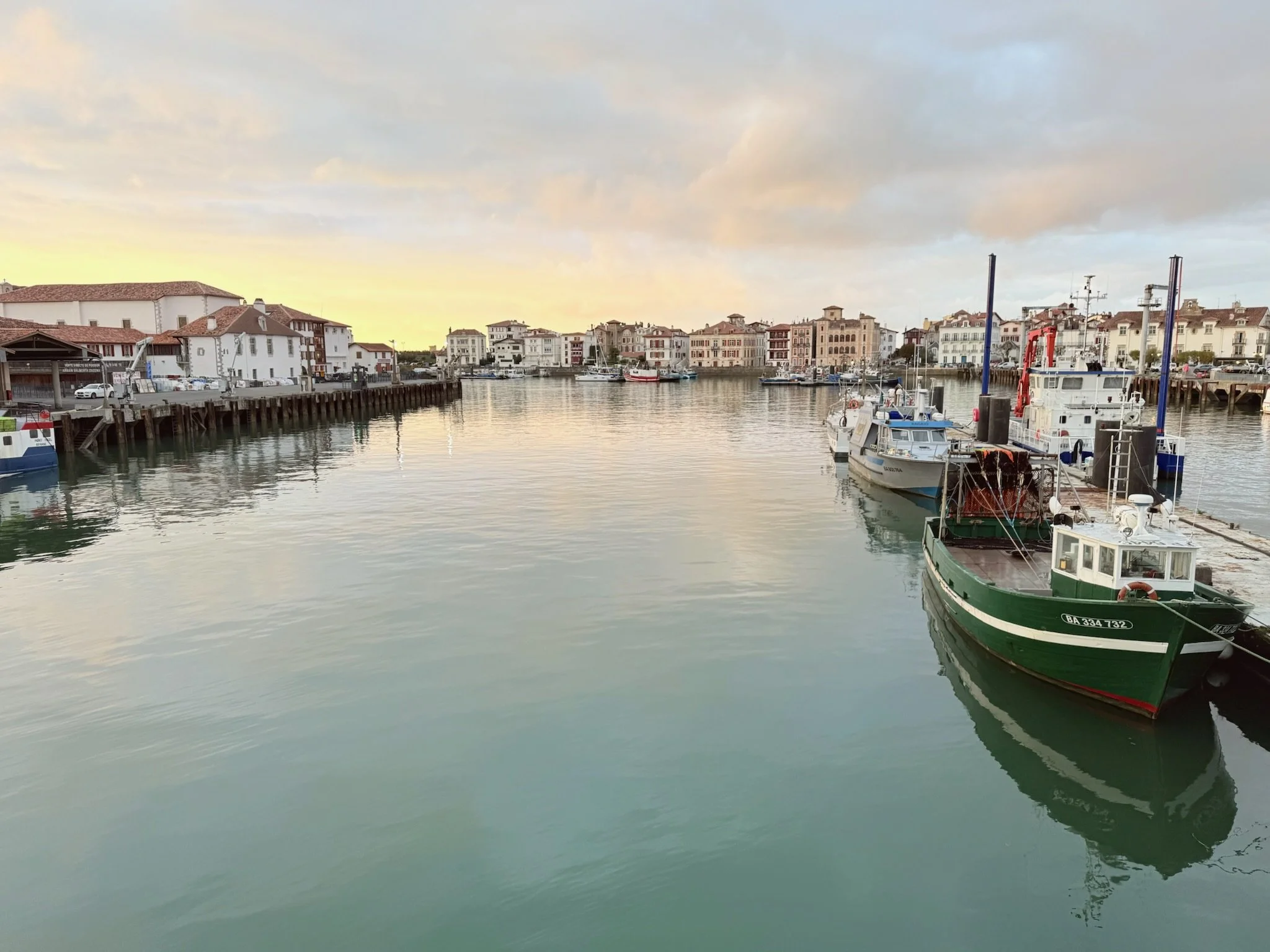 A marina with boats docked along the pier and multi-story residential buildings in the background during sunset. Calm water reflects the boats and buildings.