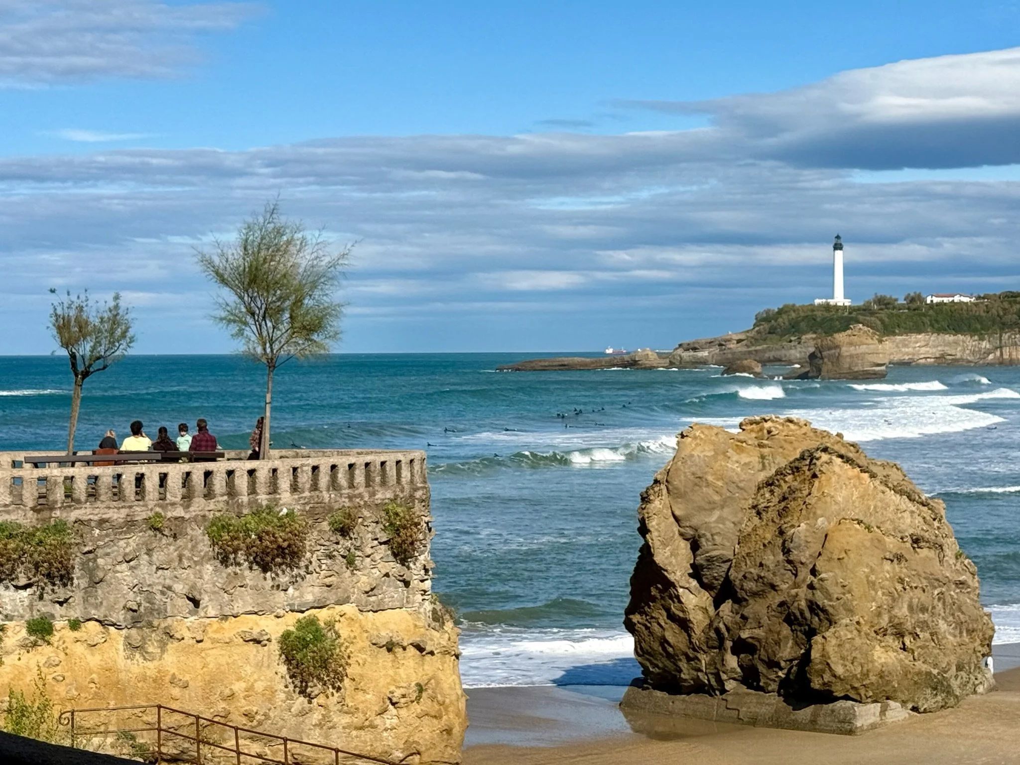 People sitting on a bench near the ocean with trees and large rocks in the foreground, a lighthouse on a hill in the background, under a partly cloudy sky.