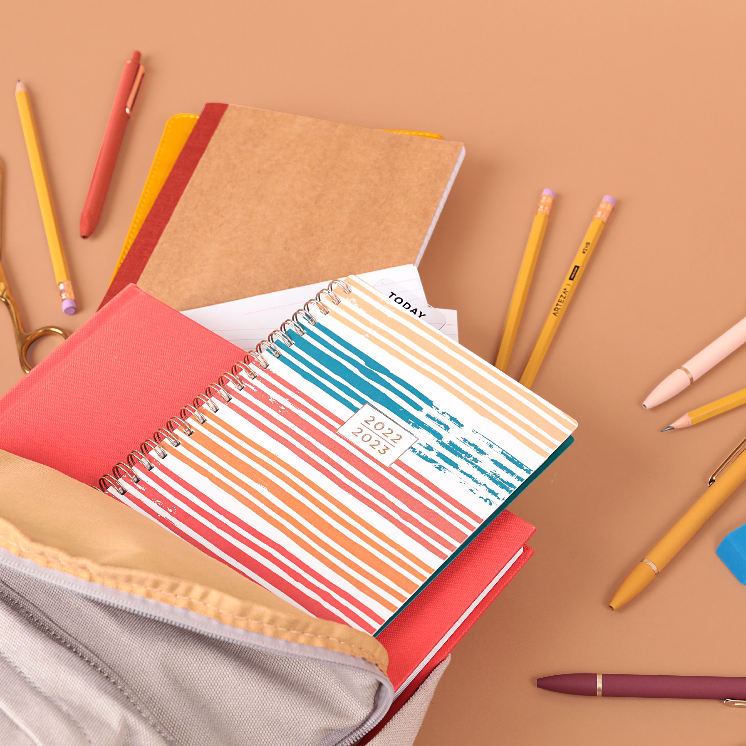 A desk with colorful notebooks, including a 2022-2023 planner, surrounded by yellow pencils, a pink pen, a purple pen, and a beige zippered pouch on a peach-colored background.