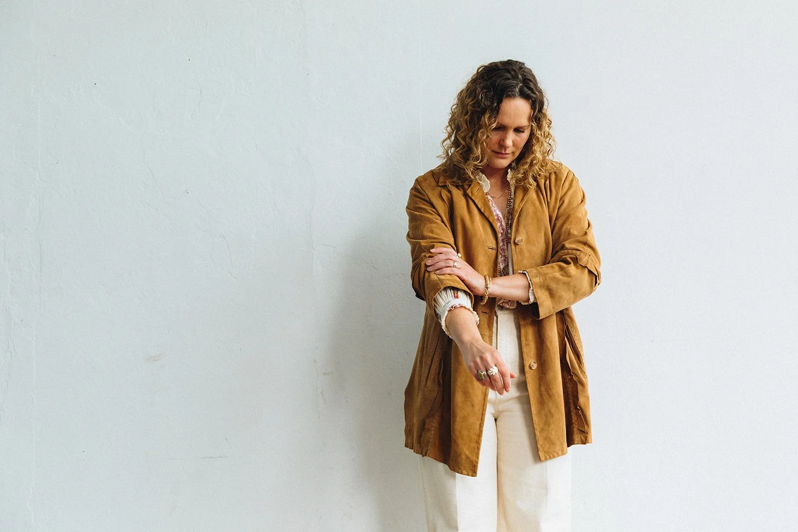 A woman with curly hair wearing a tan coat, white shirt, and light-colored pants standing with her arms crossed against a plain white wall.