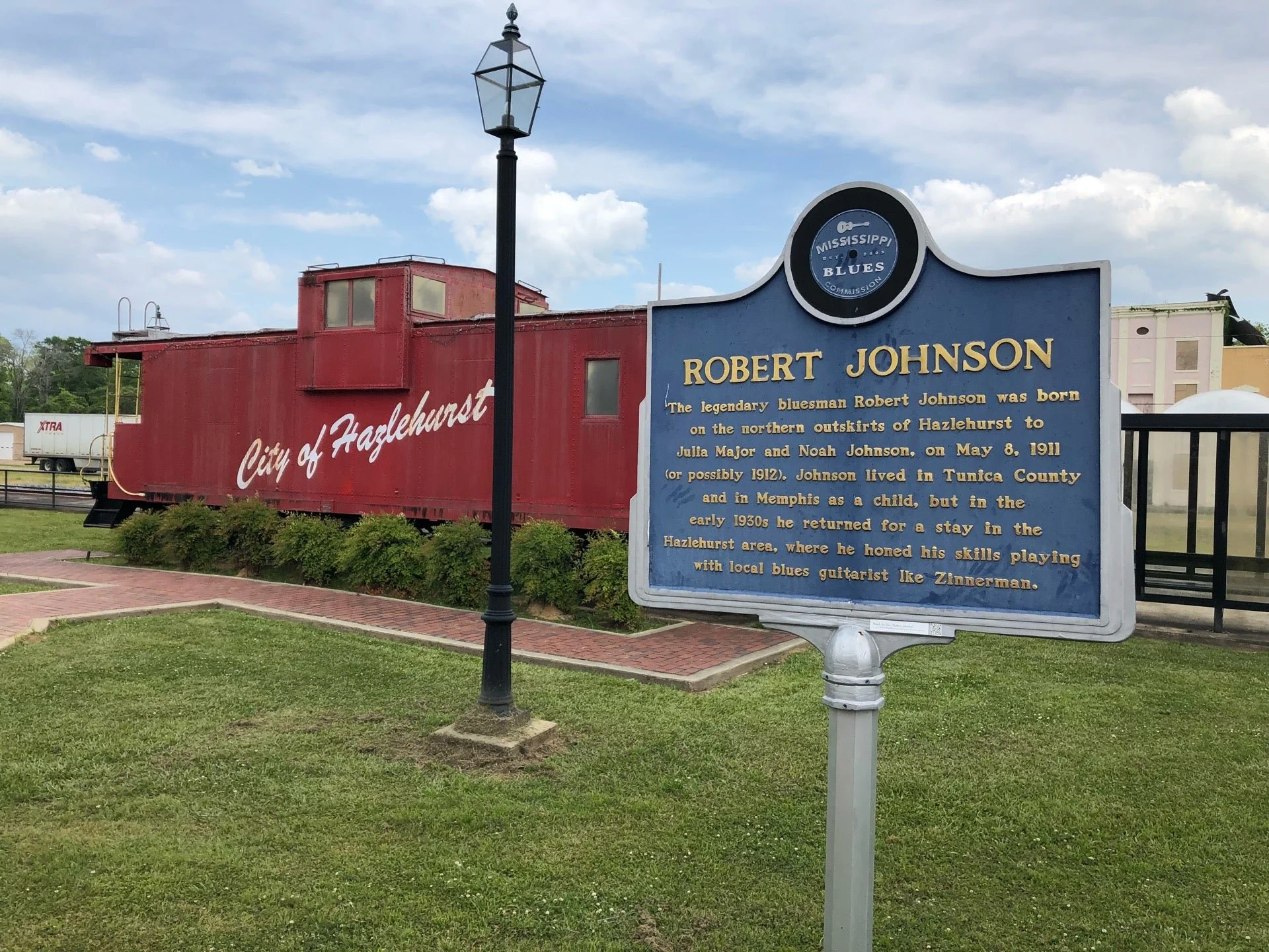 Red train car labeled 'City of Hazlehurst' next to a historical marker about blues musician Robert Johnson, located in Hazlehurst, Mississippi, under a cloudy sky.