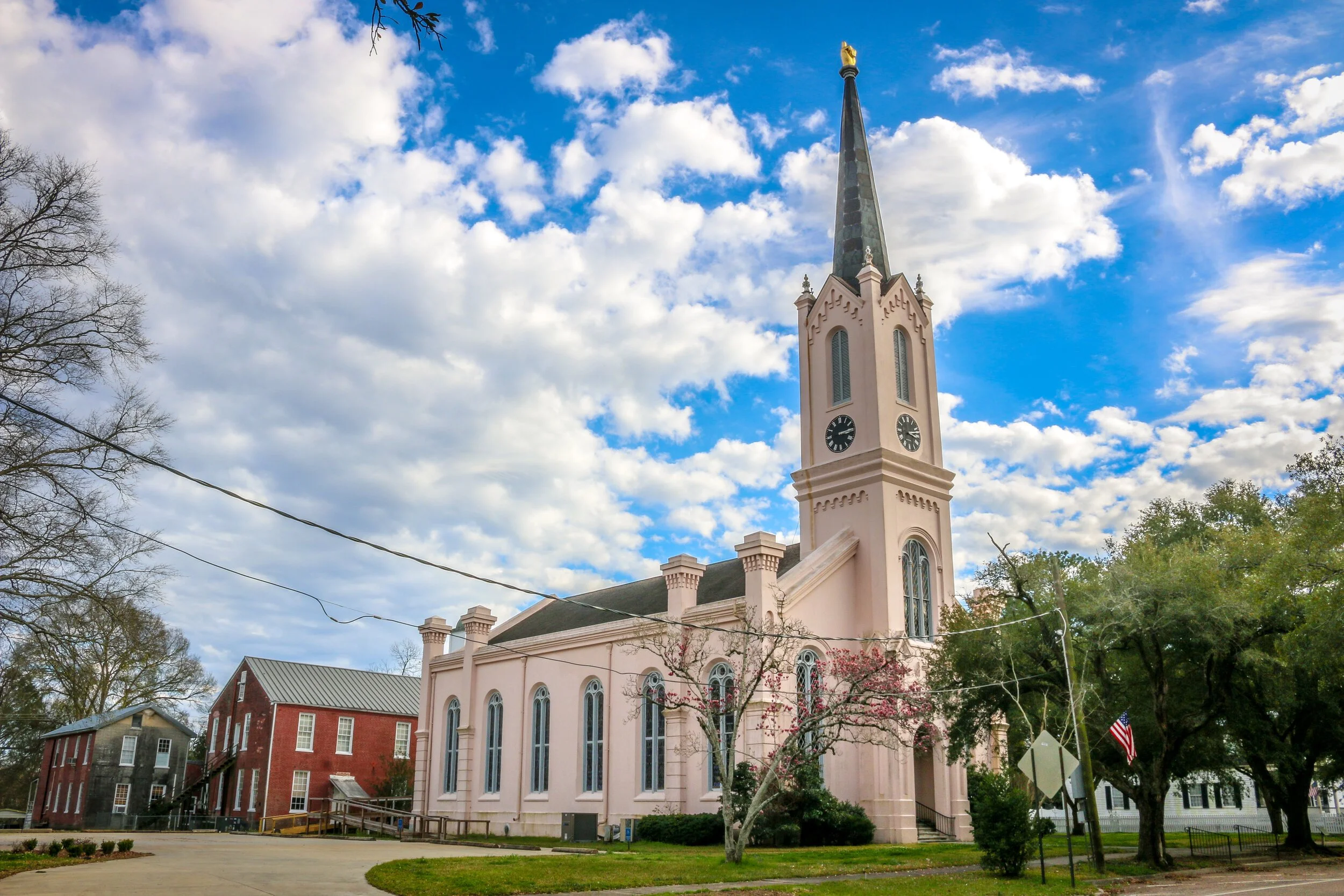 Historic church with clock tower and steeple, surrounded by trees and old brick buildings, under partly cloudy sky.