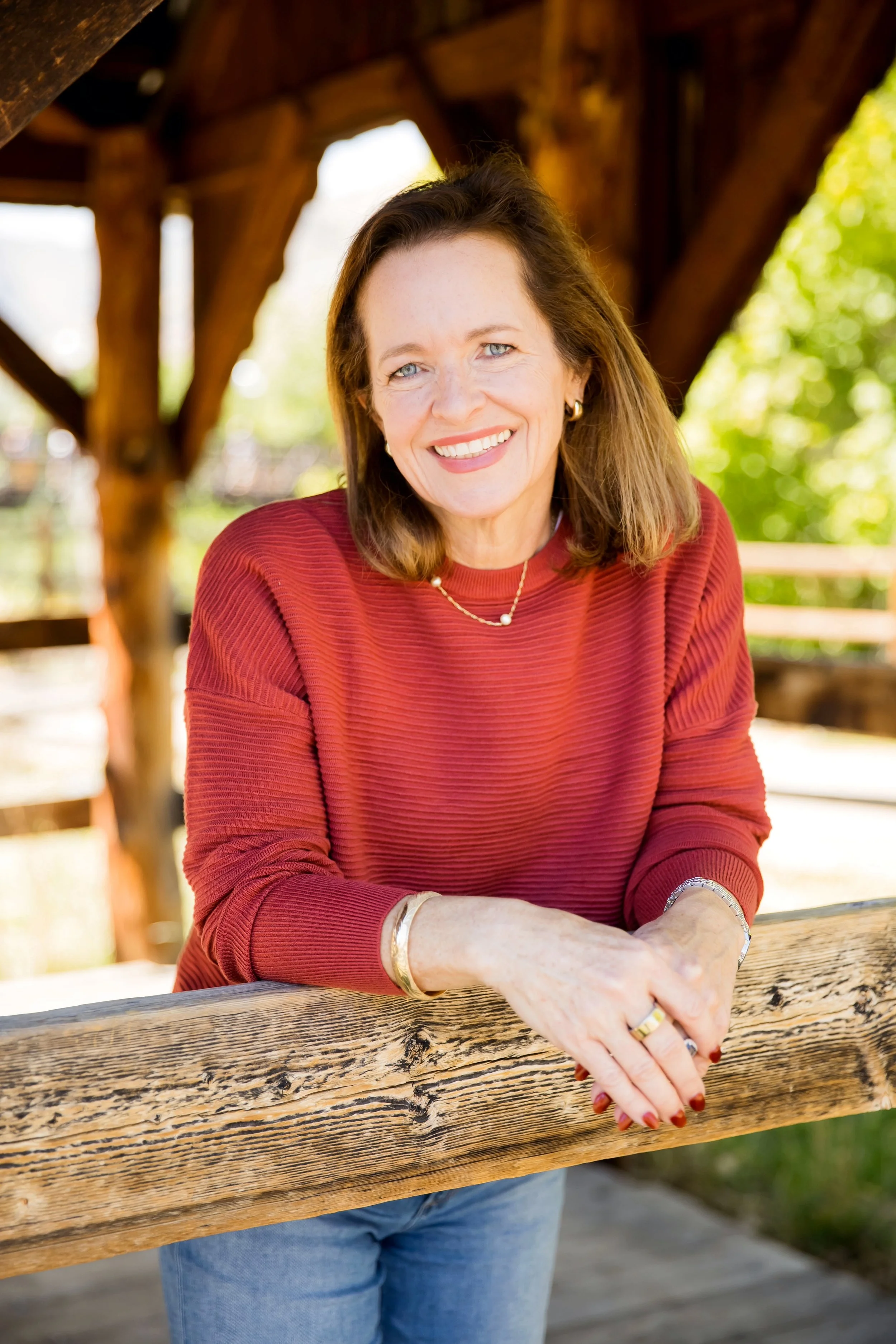 A smiling woman with shoulder-length brown hair, wearing a red long-sleeve shirt and jewelry, poses outdoors on a wooden deck with a rustic wooden structure in the background and green trees.