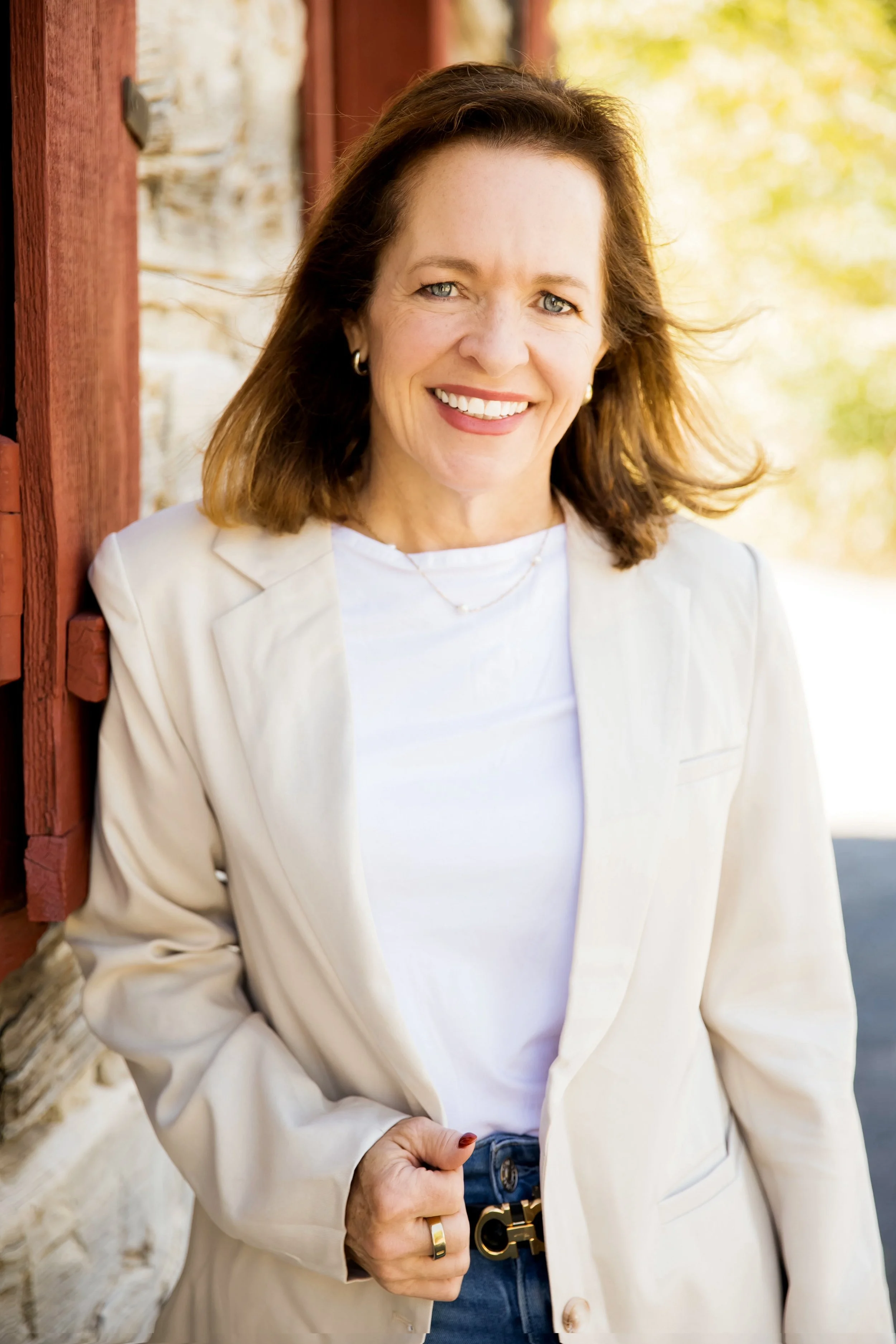 Woman with shoulder-length brown hair, wearing a white blazer over a white top, with blue jeans and a black belt, smiling and leaning against a red-painted wooden wall.